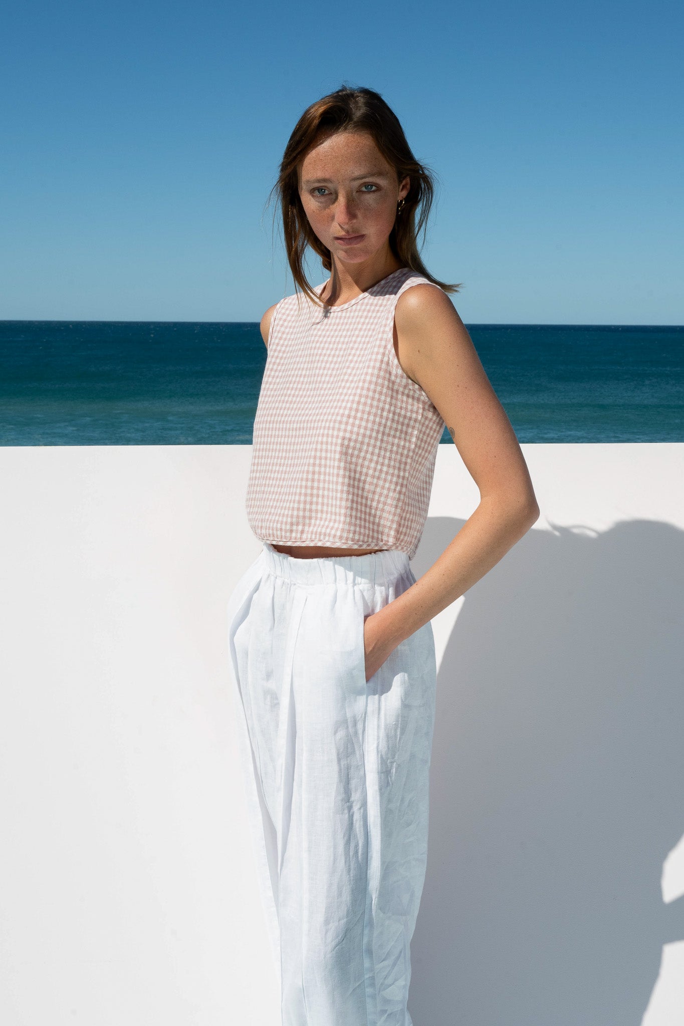 Angled view of The Boy & I Box Crop Top in blush gingham with ocean backdrop