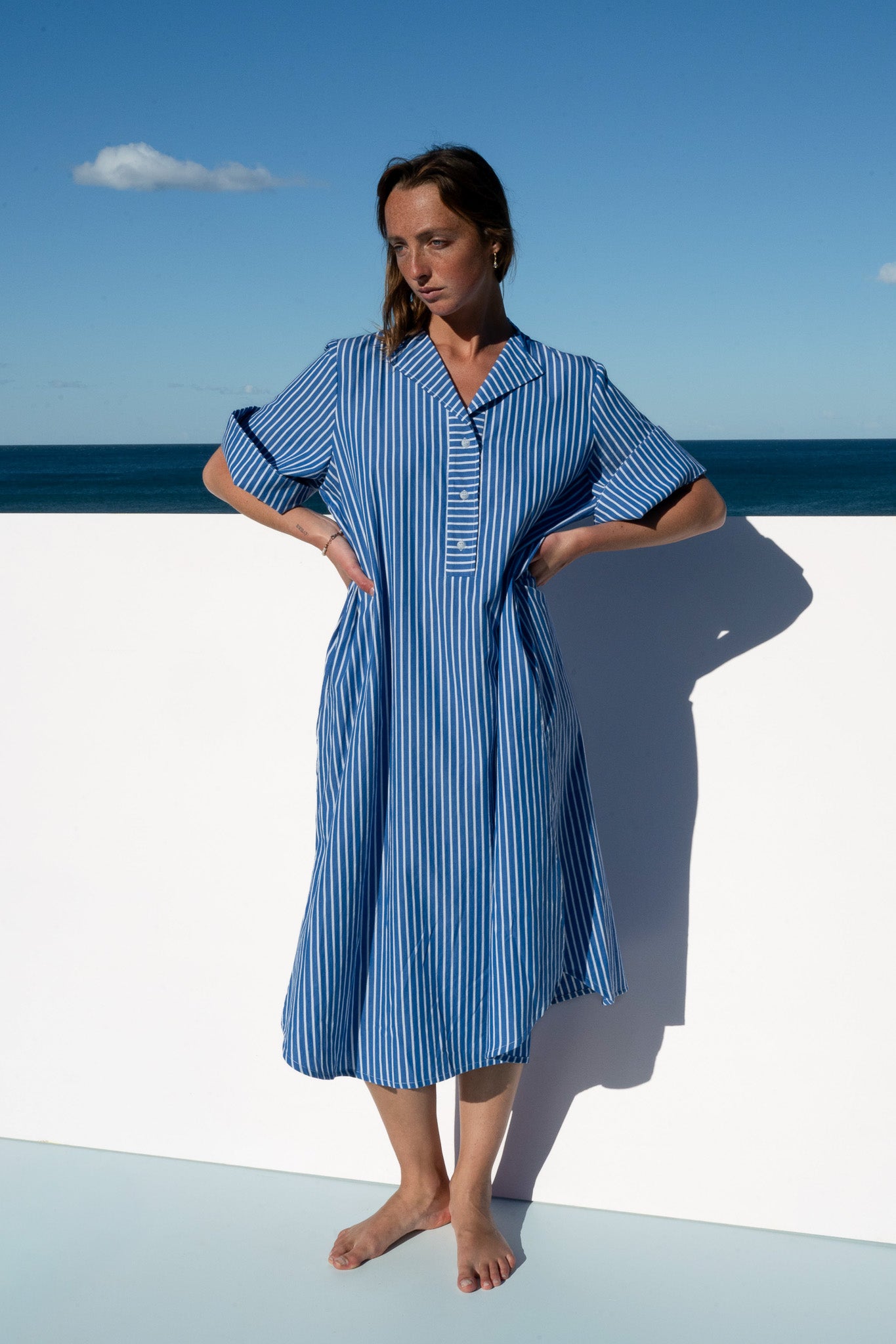 Model wearing the Cabana Dress in blue stripe cotton, standing with hands on waist against a coastal backdrop.