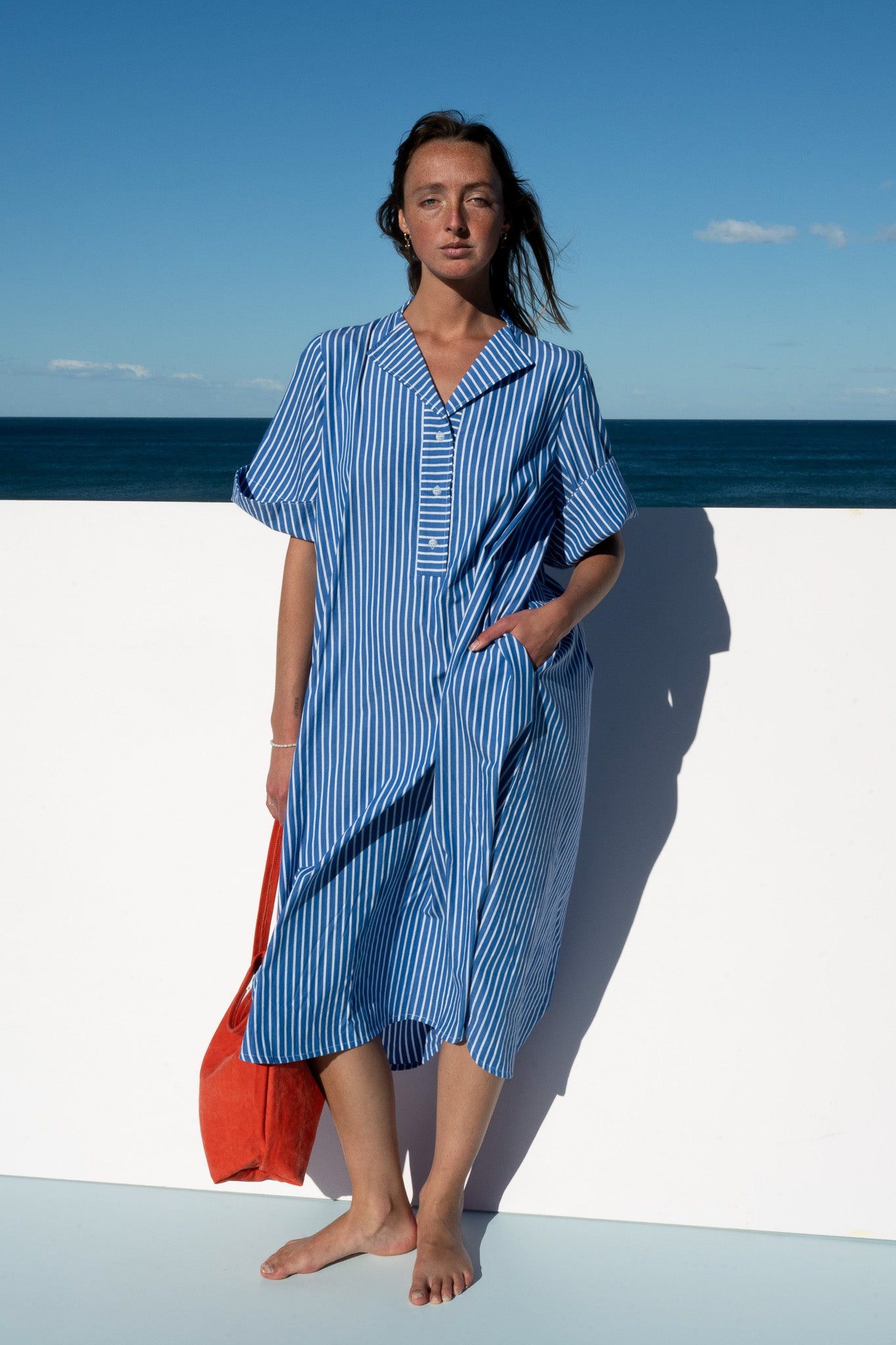 Model wearing the Cabana Dress in blue stripe cotton, hands in pockets with an orange tote by her side.