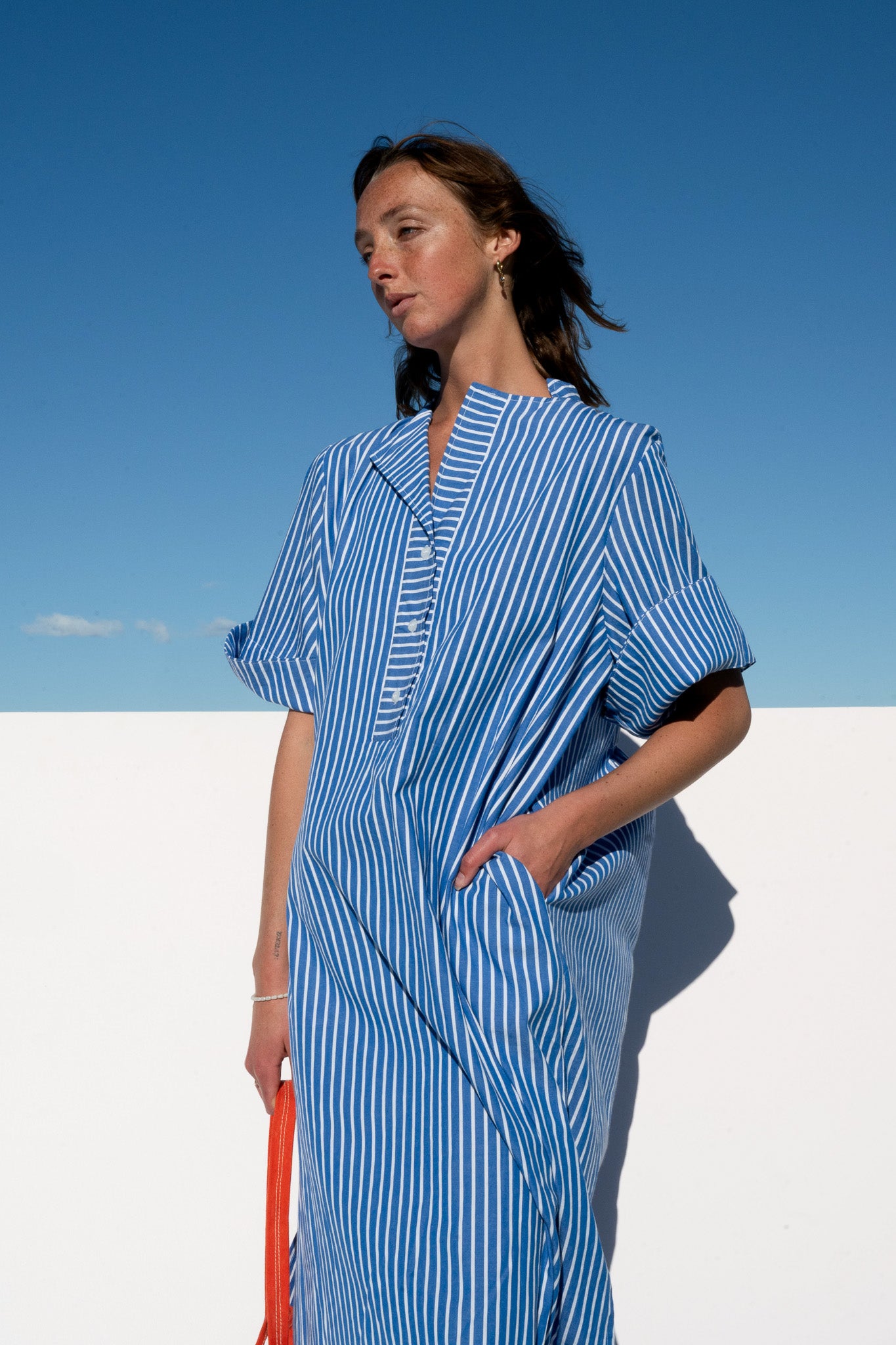 Model wearing the Cabana Dress in blue stripe cotton, front side view with hands in pocket.