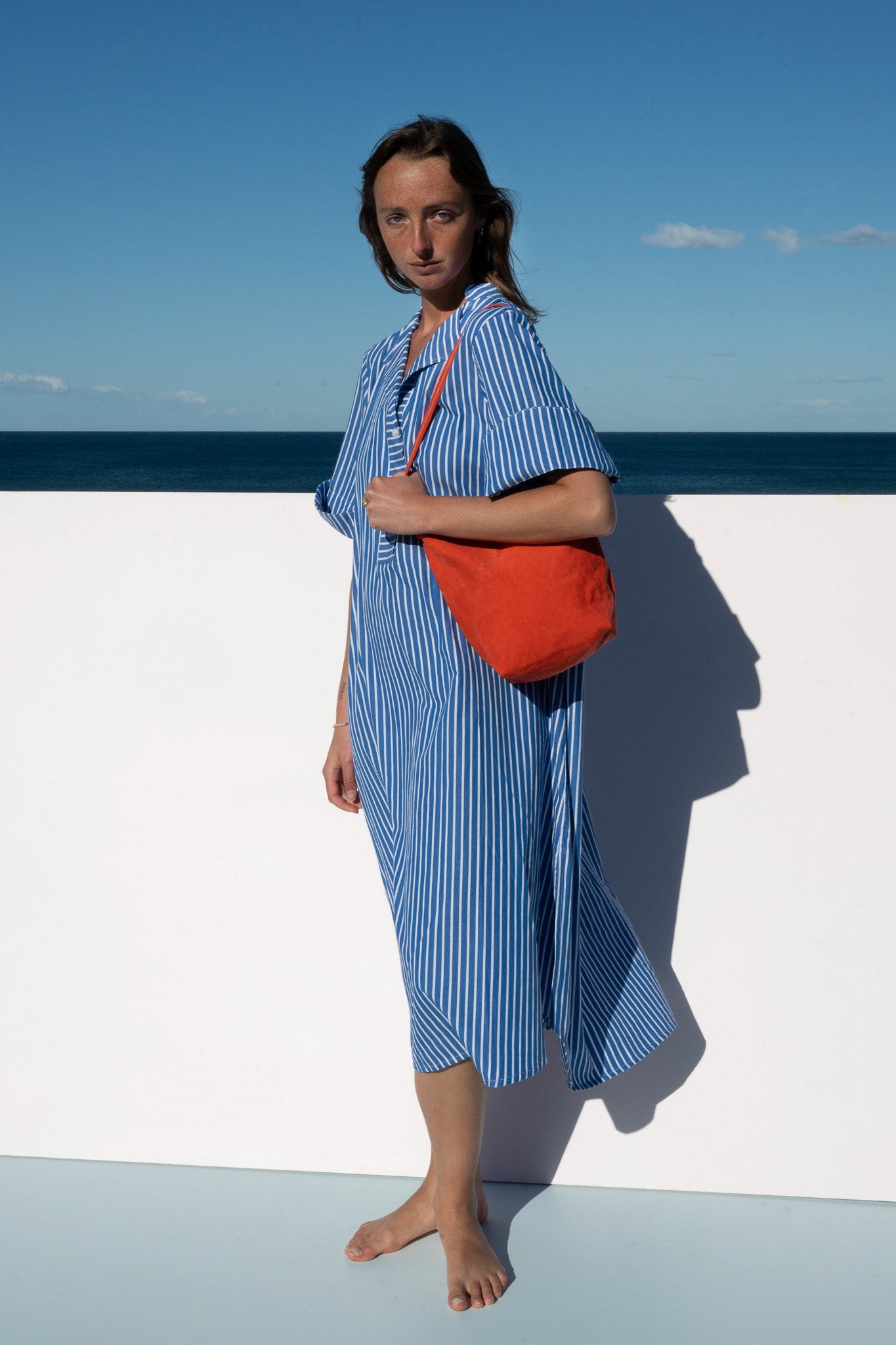 Side view of the Cabana Dress in blue stripe cotton, styled with a bold orange shoulder bag.