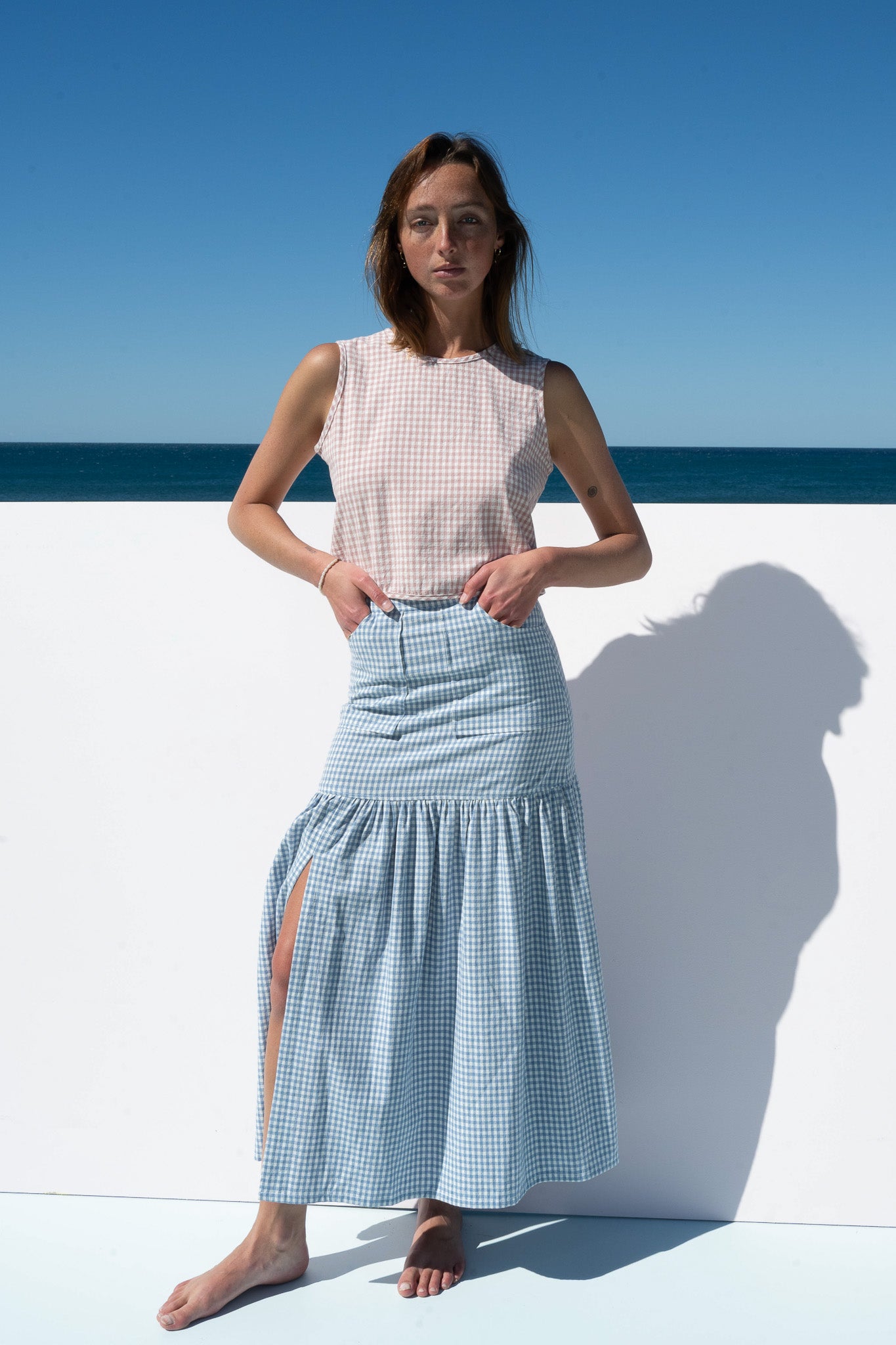 Full-length front view of Cancan Skirt blue gingham with blush gingham crop top, styled barefoot against ocean backdrop.