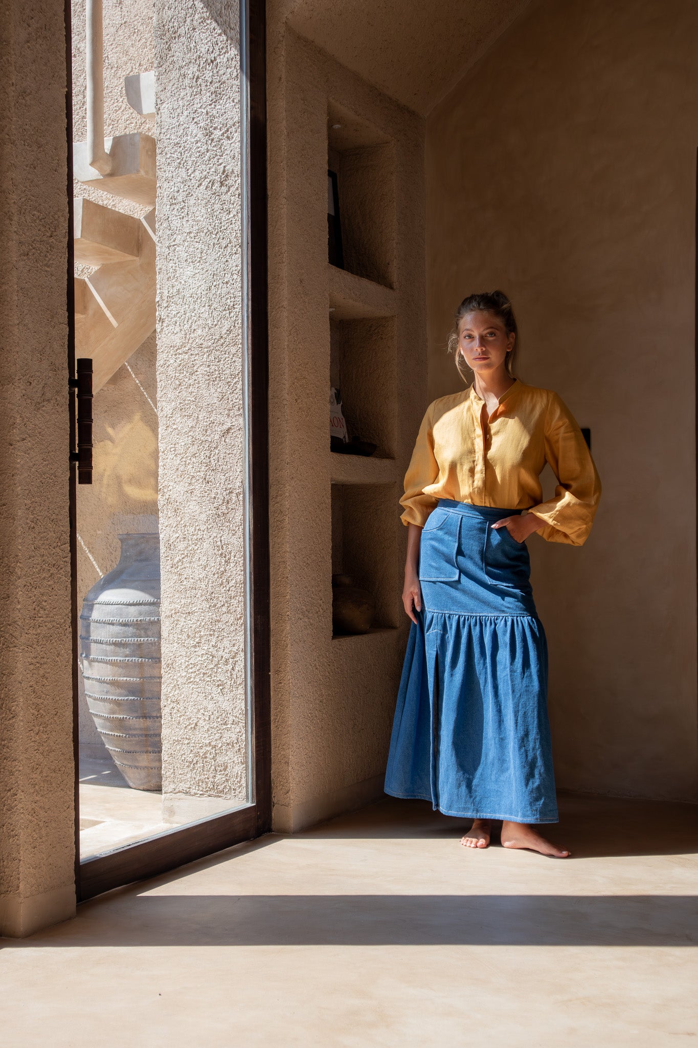 Model in Coconut Blouse in mango and denim skirt standing barefoot in natural light, with minimalist decor in the background.