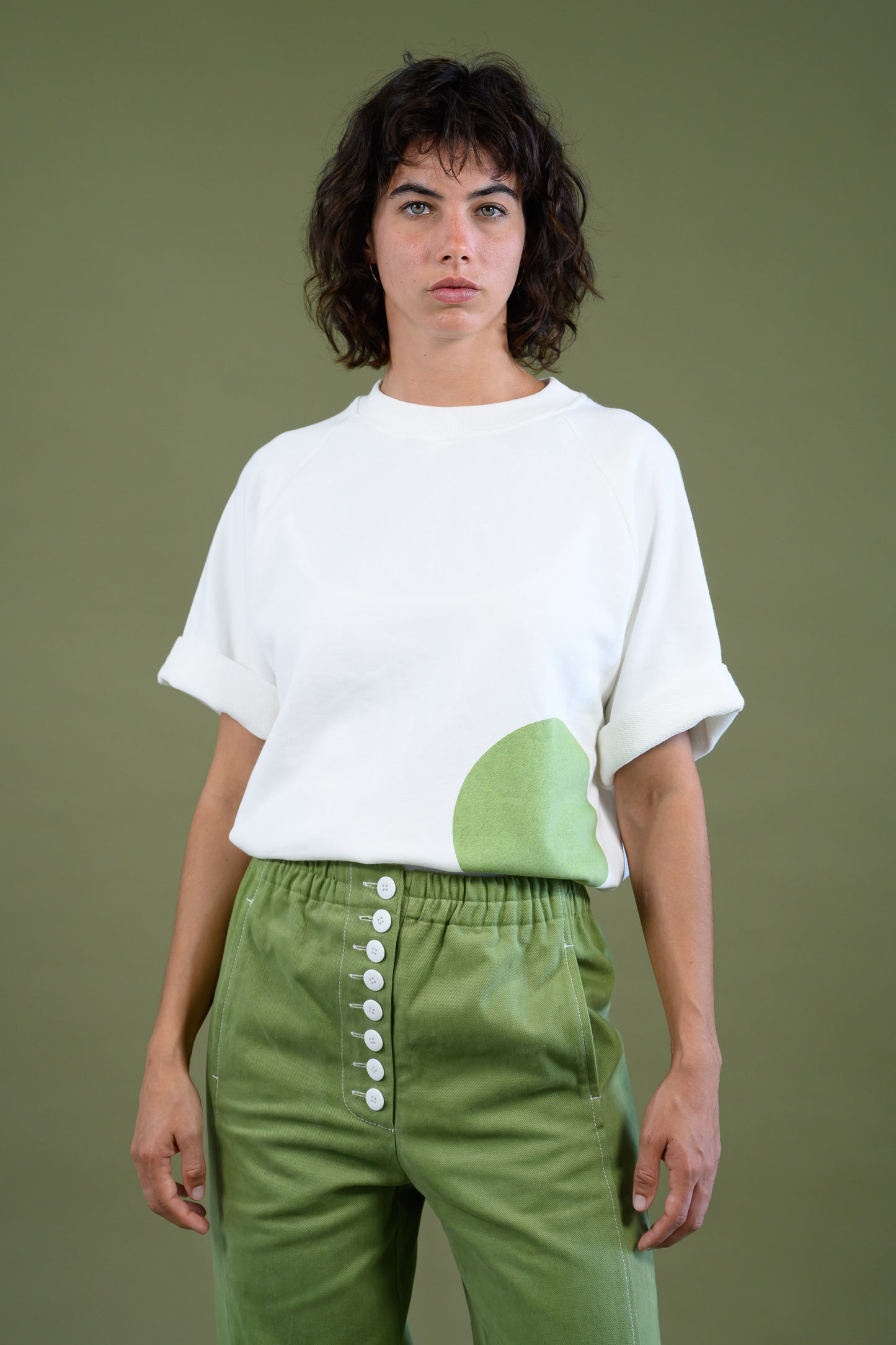 Model facing the camera wearing white sweatshirt with raglan sleeves and green dot feature.