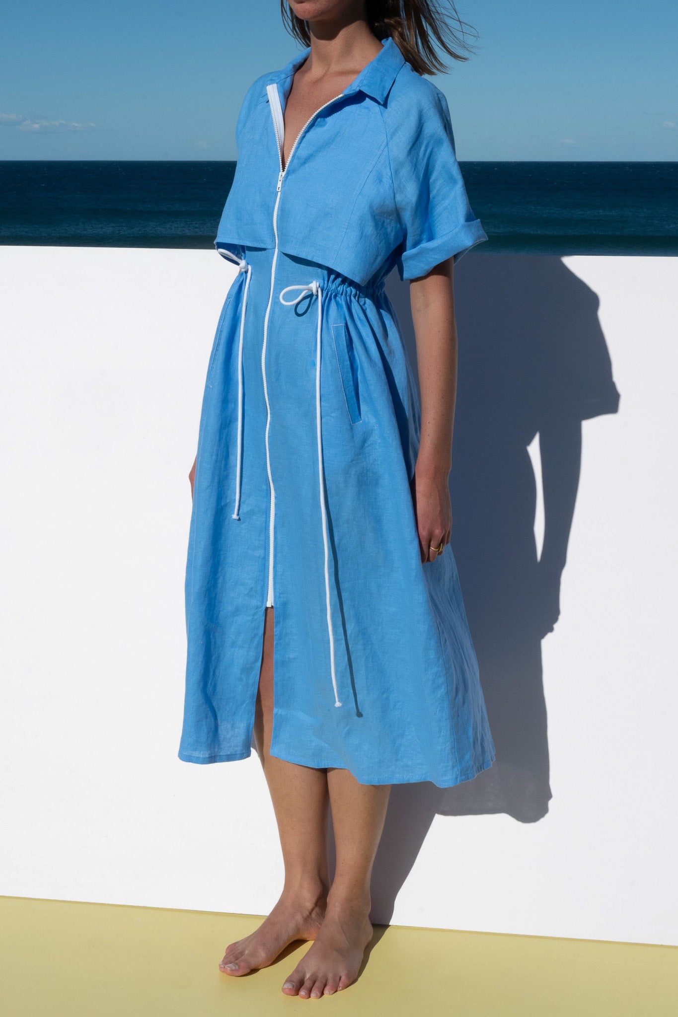Half-length view of The Boy and I Iconica Dress in blue linen with white zip and drawstrings.