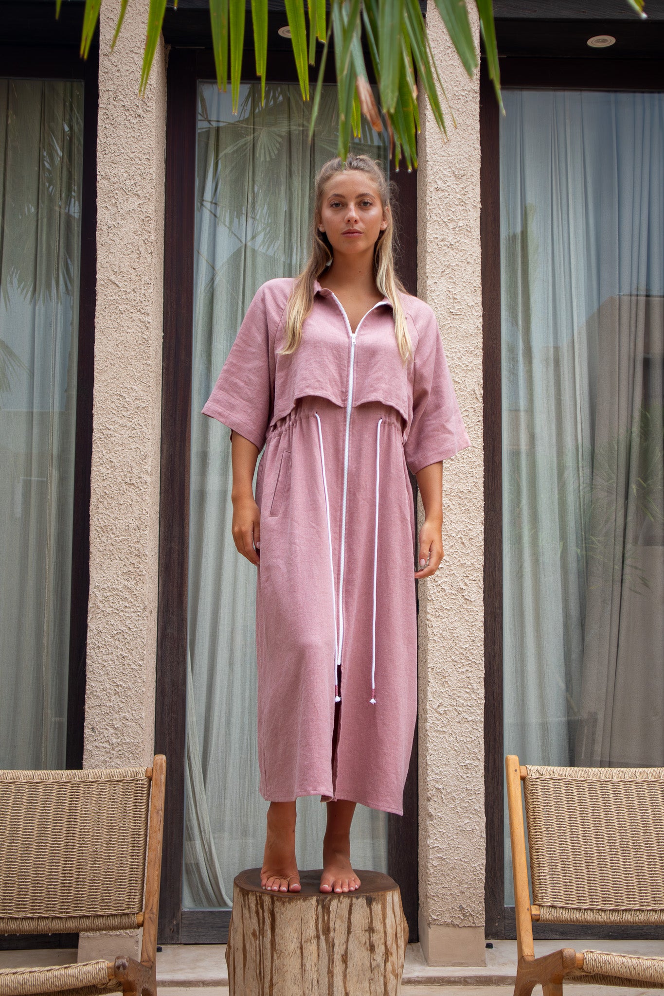 Model standing on wooden stump in full-length view of dusty pink Iconica dress with drawstring waist and zip front