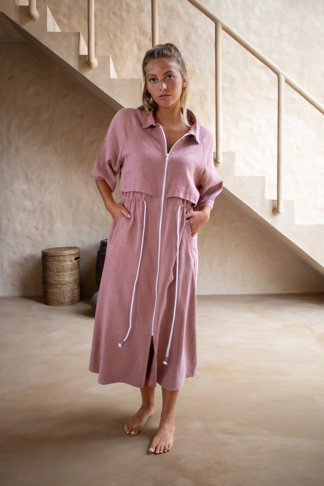 Model wearing dusty pink Iconica dress with cropped overlay and gathered waist, photographed indoors with soft natural light