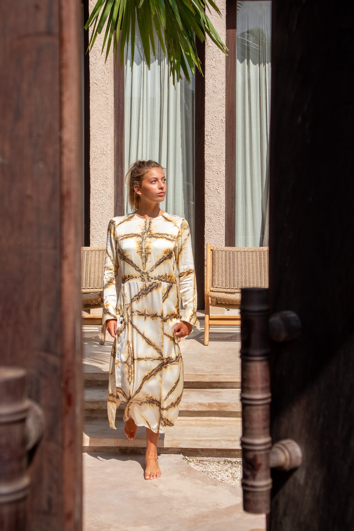 Model stepping barefoot through wooden doorway in Maggie Dress with Earth Shibori pattern, sunlight casting soft shadows