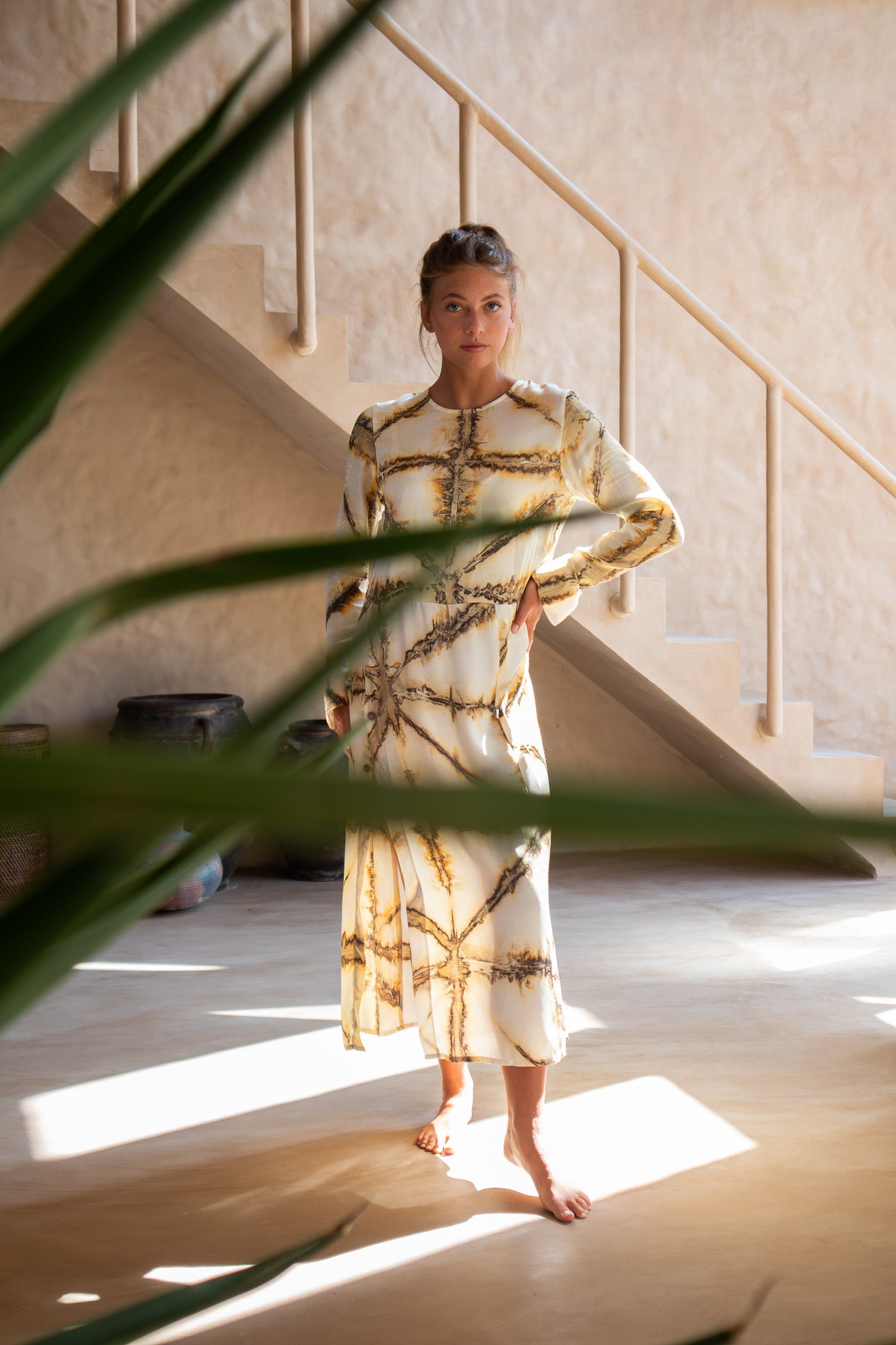 Model in Earth Shibori Maggie Dress bathed in soft sunlight with staircase and tropical leaves in foreground