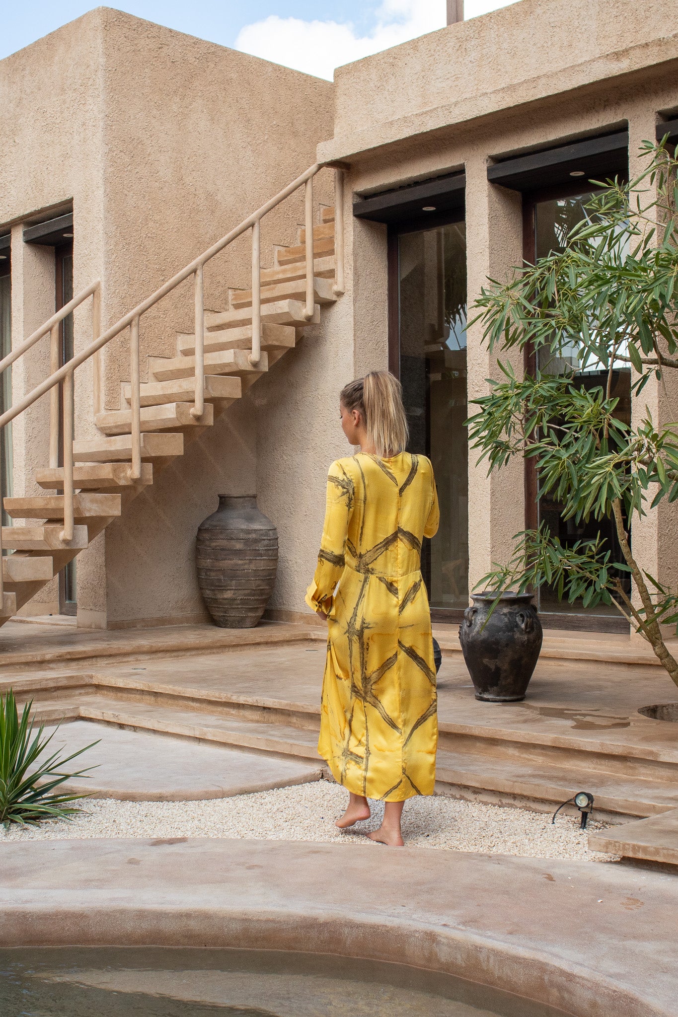 Back view of the Maggie Dress in Mango Shibori near stone steps and pool