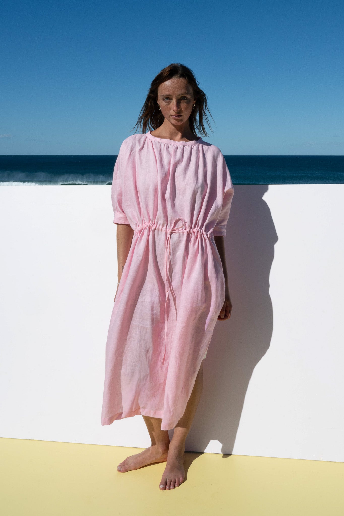 Model wearing The Moments Dress in Bonbon Pink linen, natural front view with ocean backdrop.