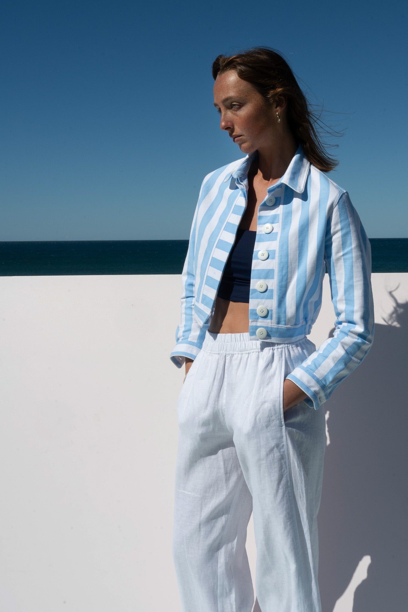 Model wearing the Solero Pant in white linen, styled with a light blue striped cropped jacket, hands in pockets, ocean backdrop.