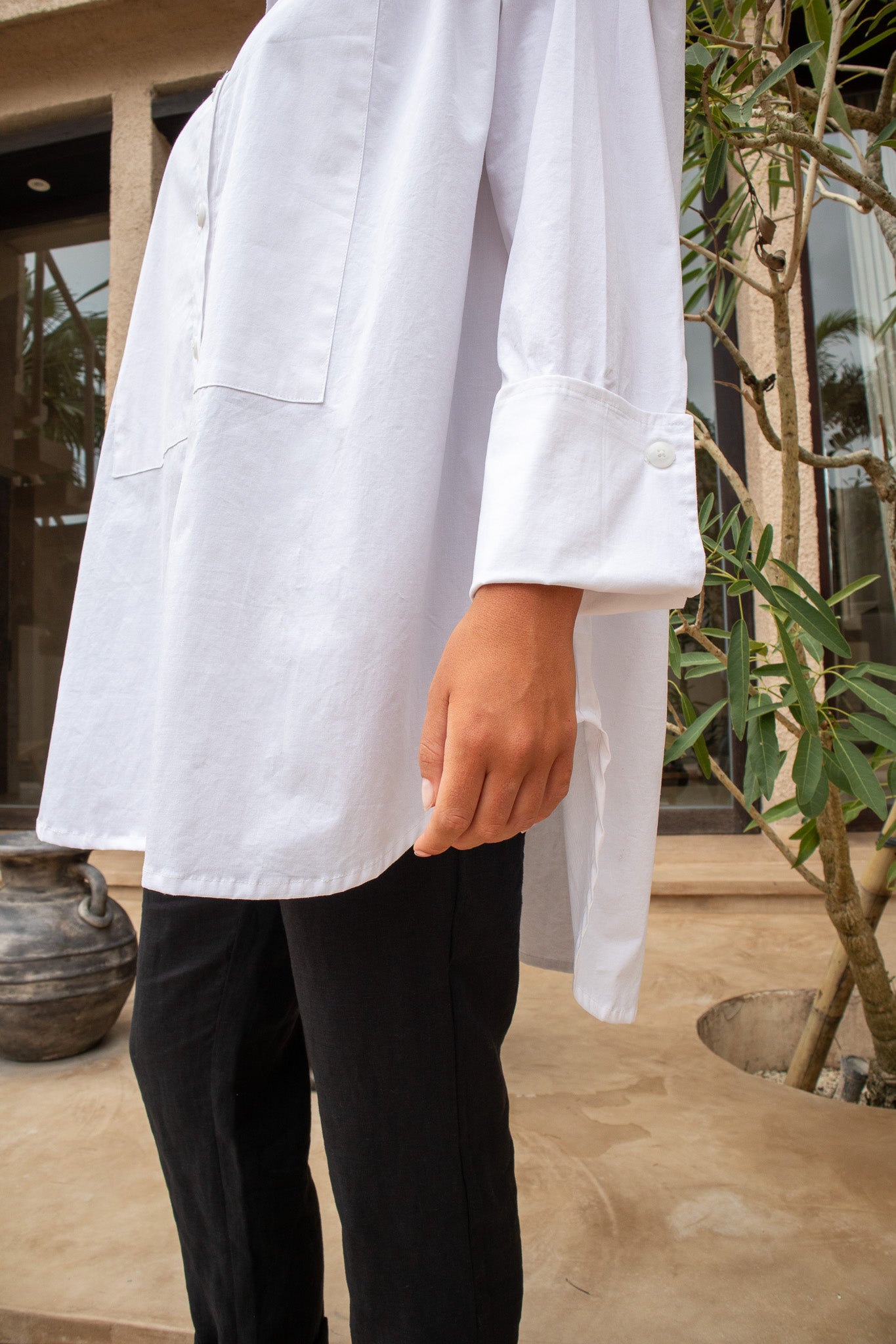 Detail of The Boyfriend Tux Shirt's cuff and side hem, styled over black pants in an outdoor garden setting.