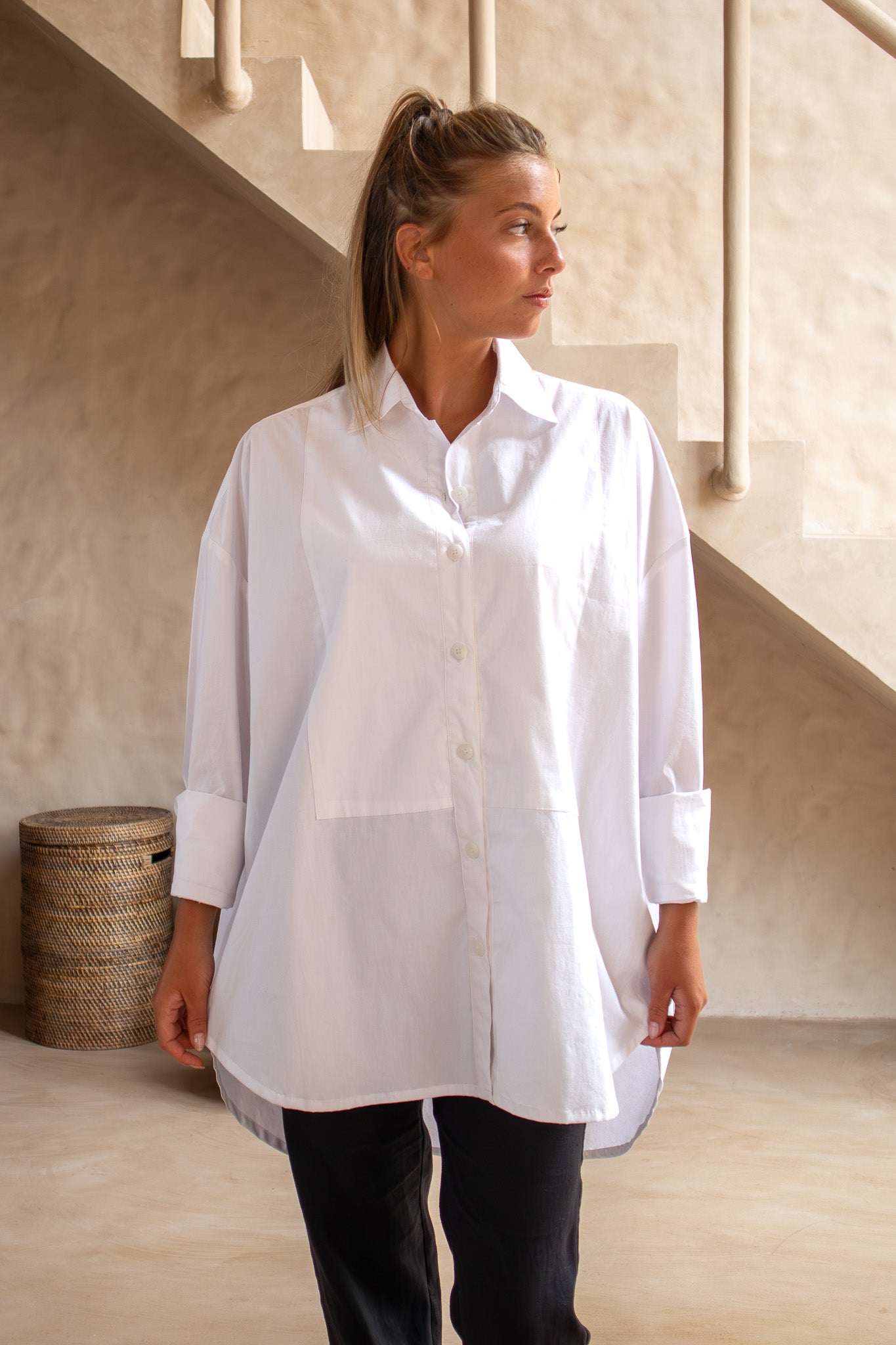 Model styling The Boyfriend Tux Shirt in white over black trousers, standing beneath a staircase and looking to the side.