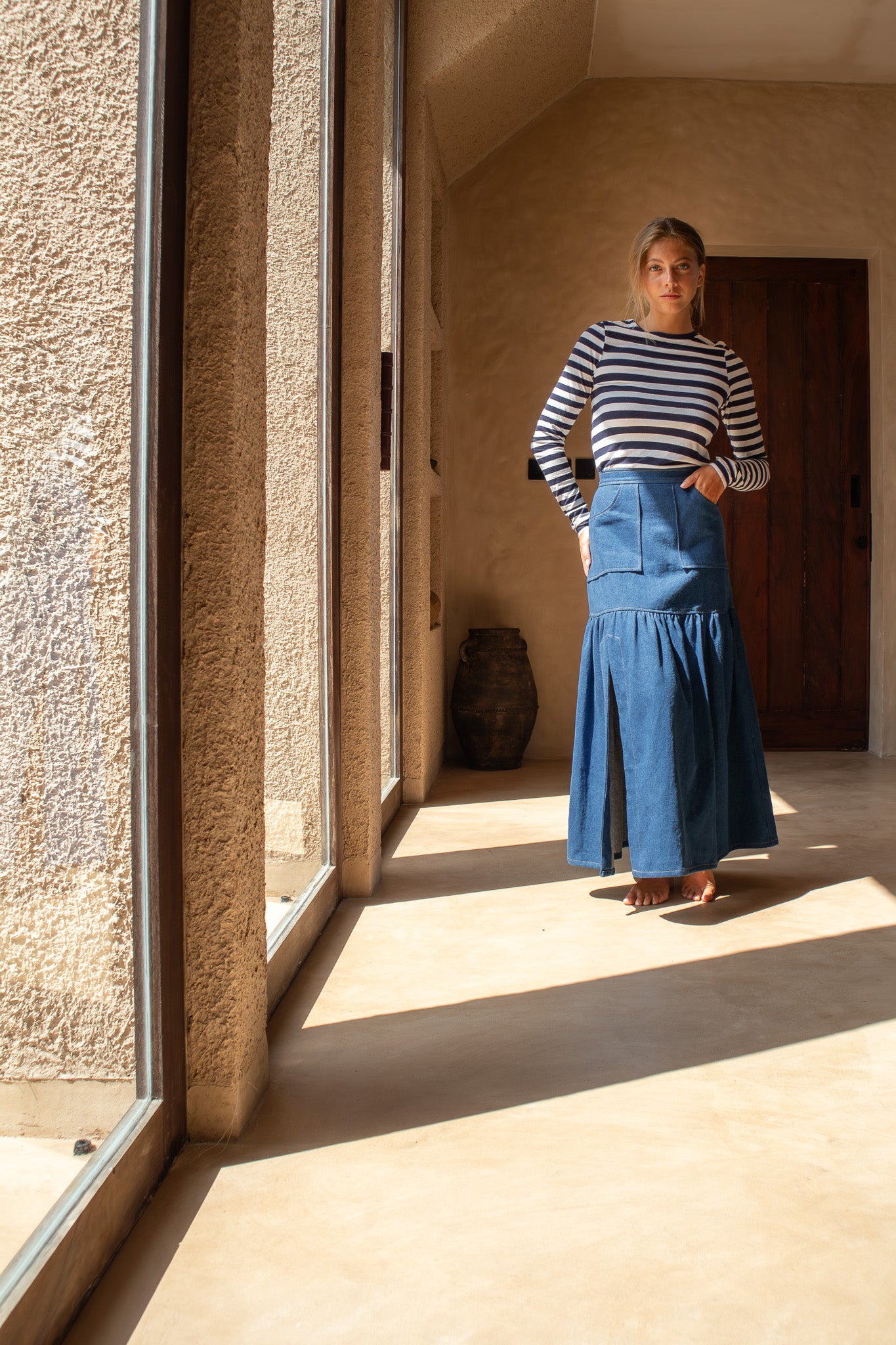 Model standing in a sunlit corridor wearing The Boy and I Cancan Skirt in denim, hands in pockets, with soft natural shadows.