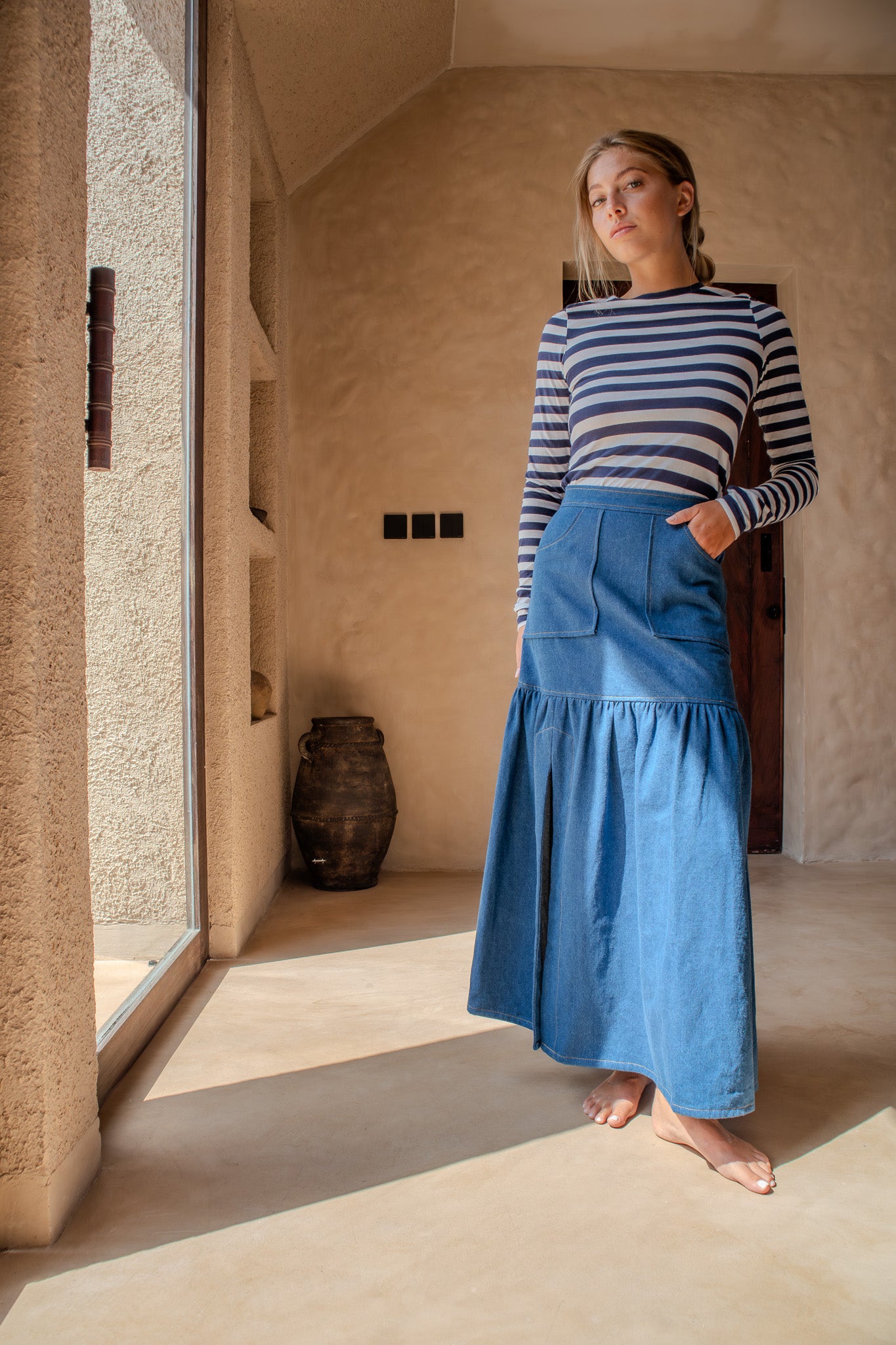 Woman in a striped top and The Boy and I Cancan Skirt in denim, standing barefoot near a window with one hand in her pocket.