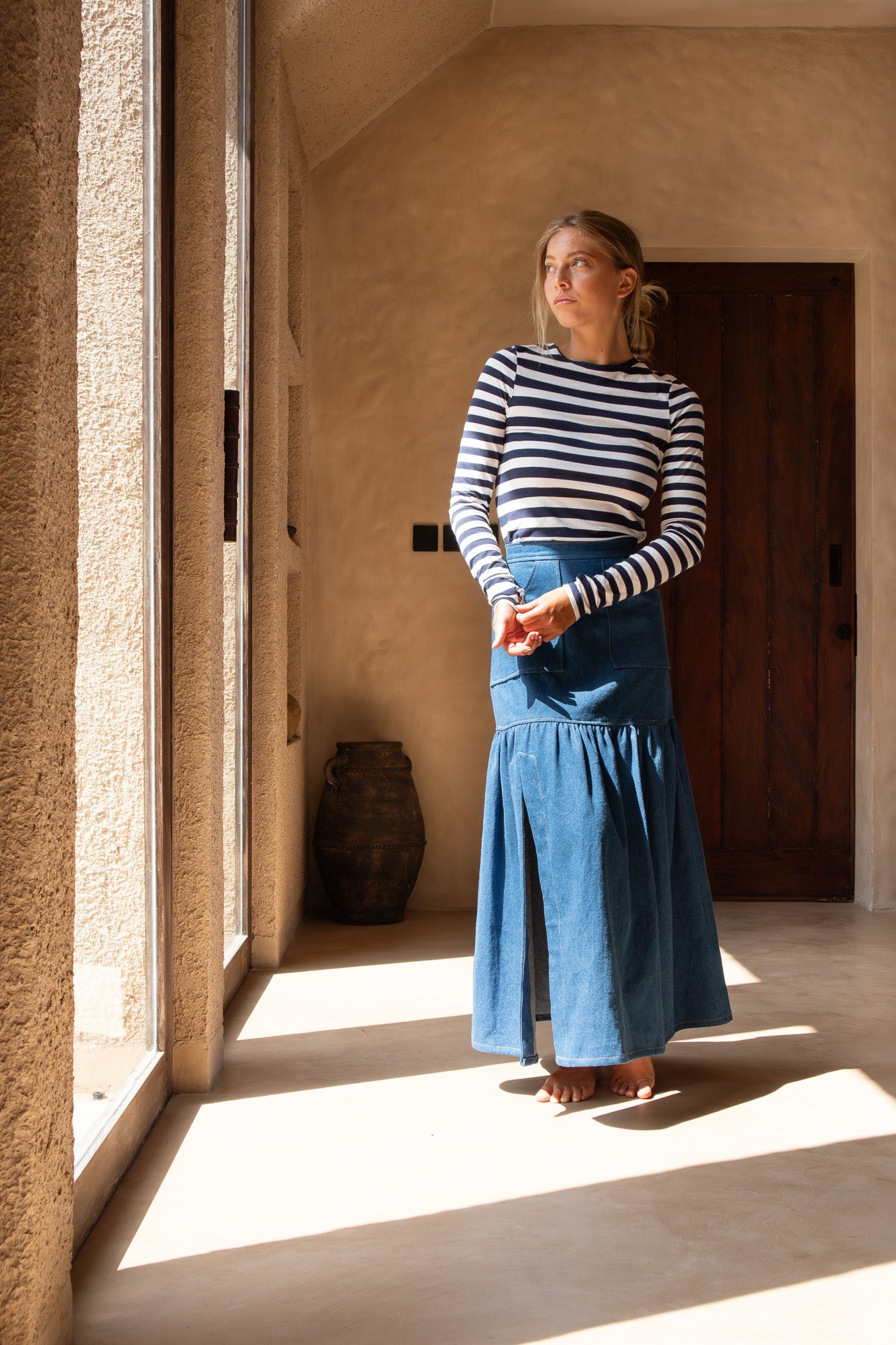 Model standing in soft light near a window, wearing The Boy and I Cancan Skirt in denim with her hands gently clasped.