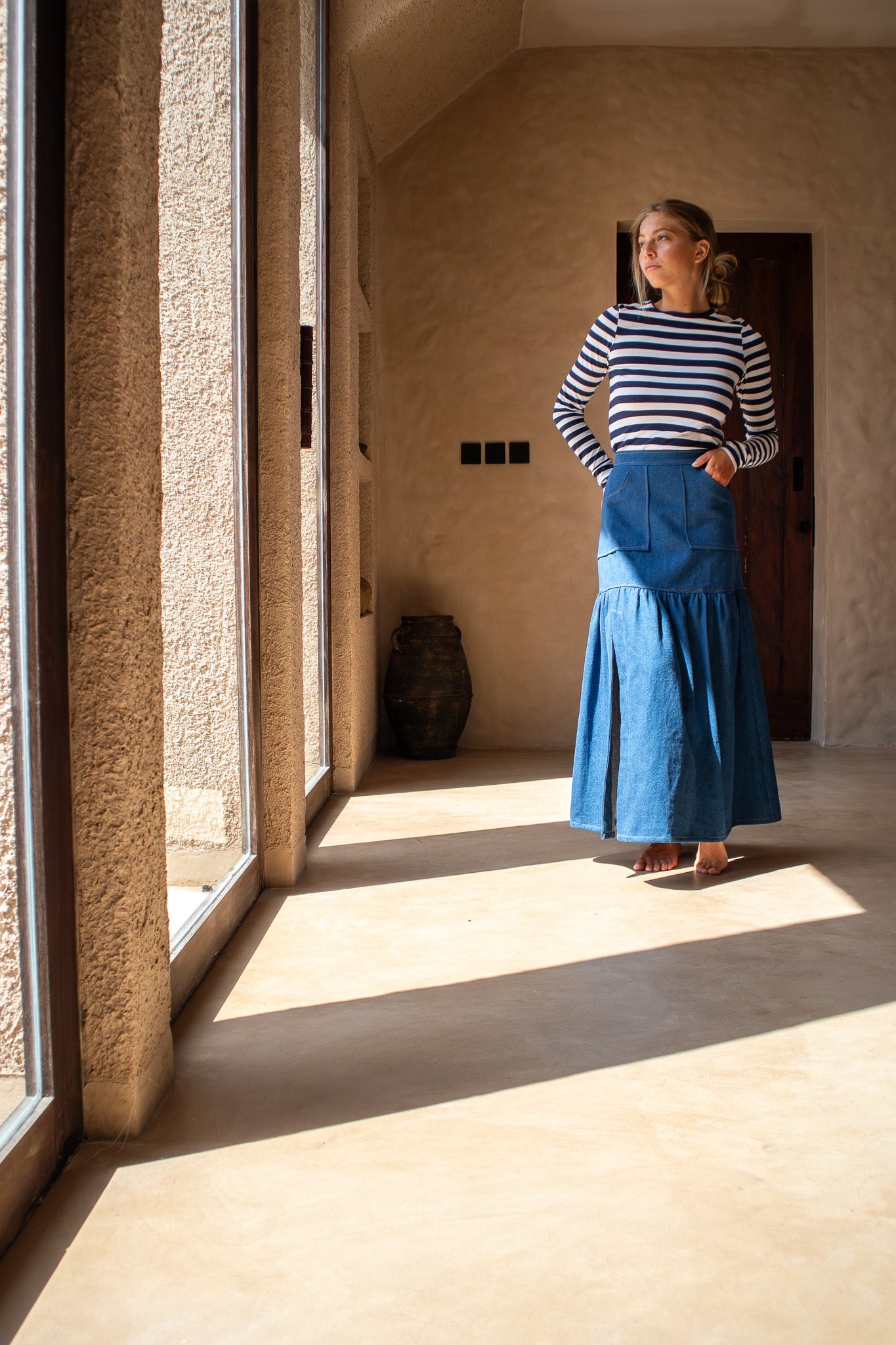 Model wearing The Boy and I Cancan Skirt in denim standing barefoot in natural window light, looking out thoughtfully.