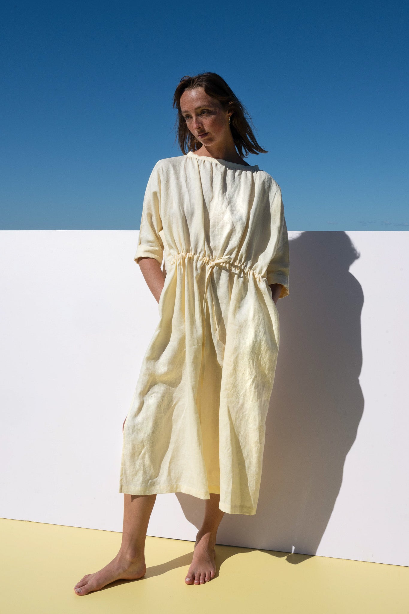 The Moments Dress in yellow linen – relaxed fit with gathered waist, model leaning in sunlight
