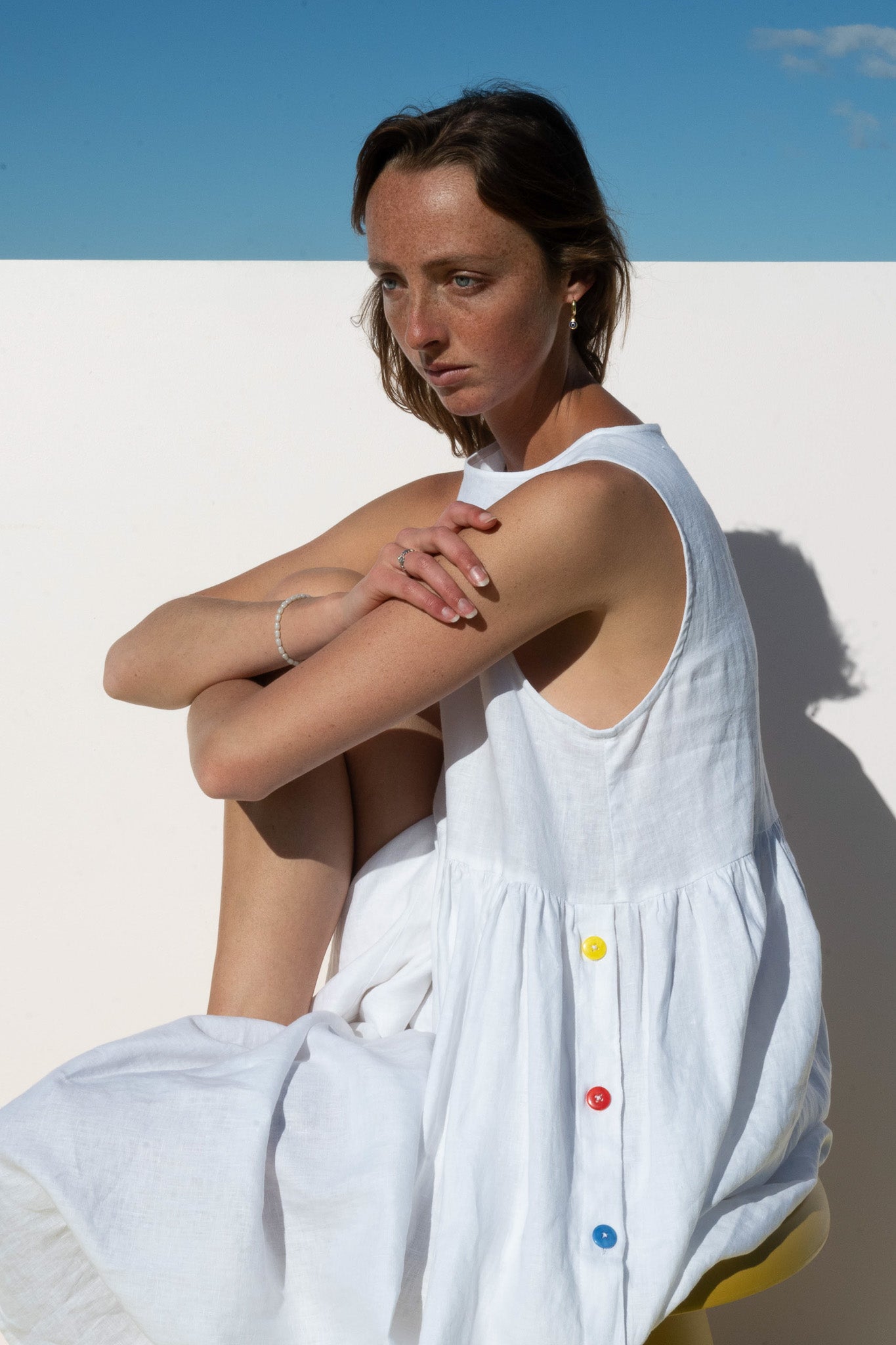 Side seated pose in the Wonka Sleeveless Dress, white linen with rainbow buttons