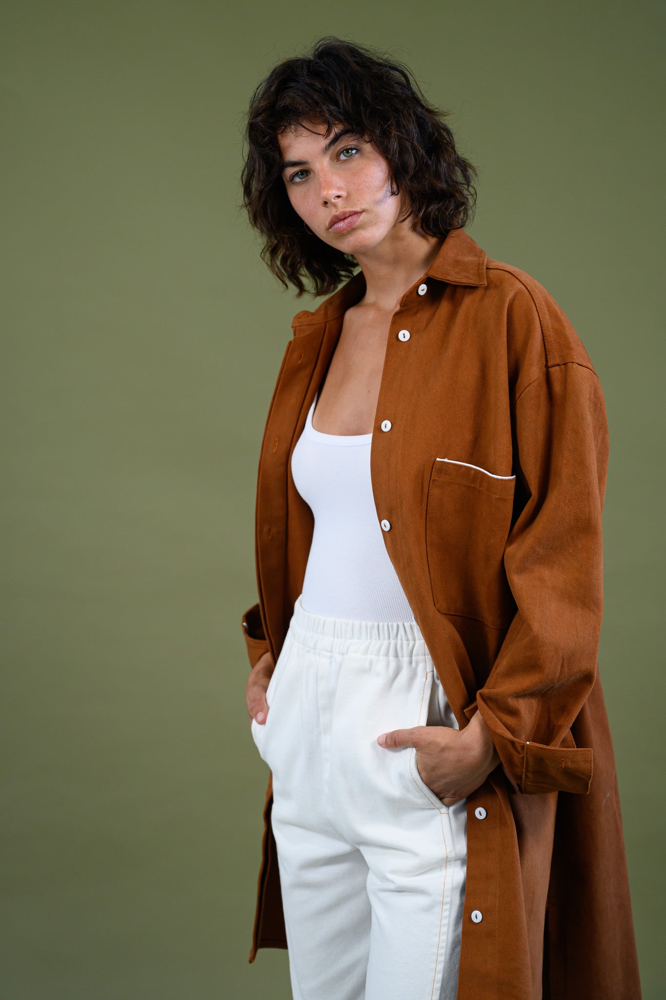 Angled portrait view of Caramel cotton canvas denim shirt coat worn open over white outfit with hands in pockets