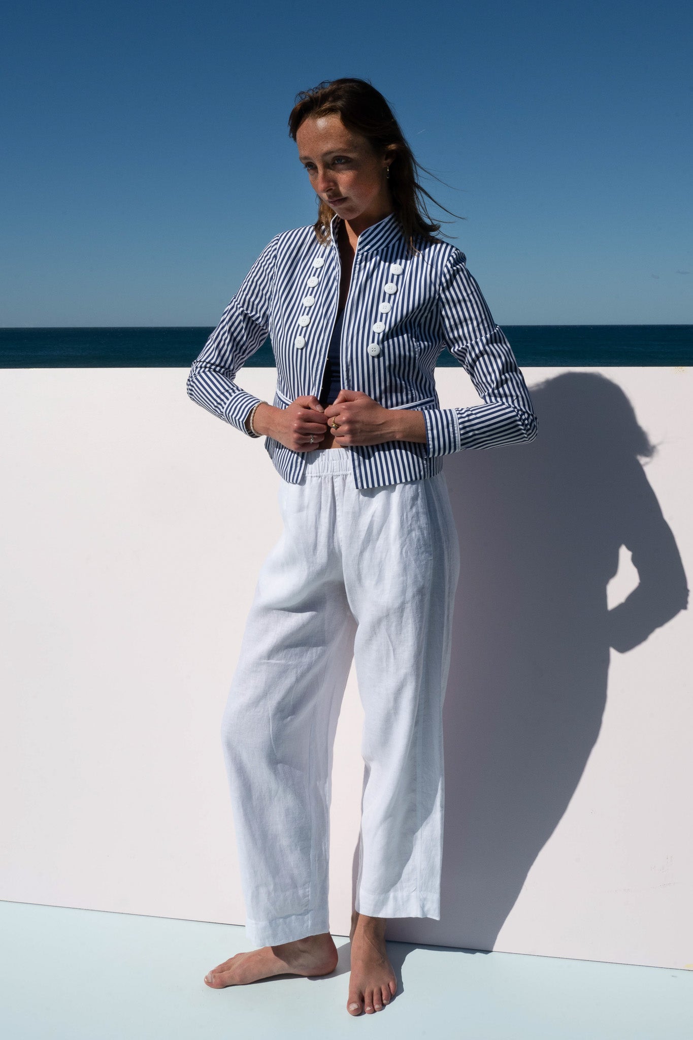 Model wearing the Abbey Road Jacket in cotton poplin azure stripe, paired with white linen pants, standing against an ocean backdrop.