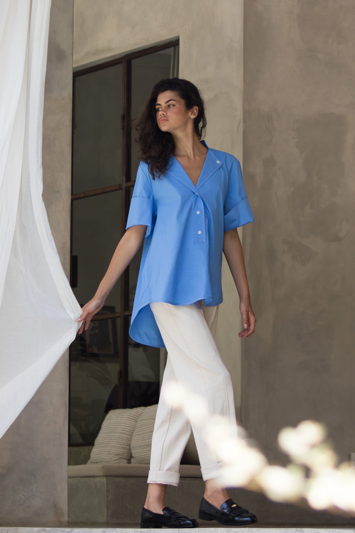 Model walking wearing blue cotton poplin blouse showing scooped hem at the back