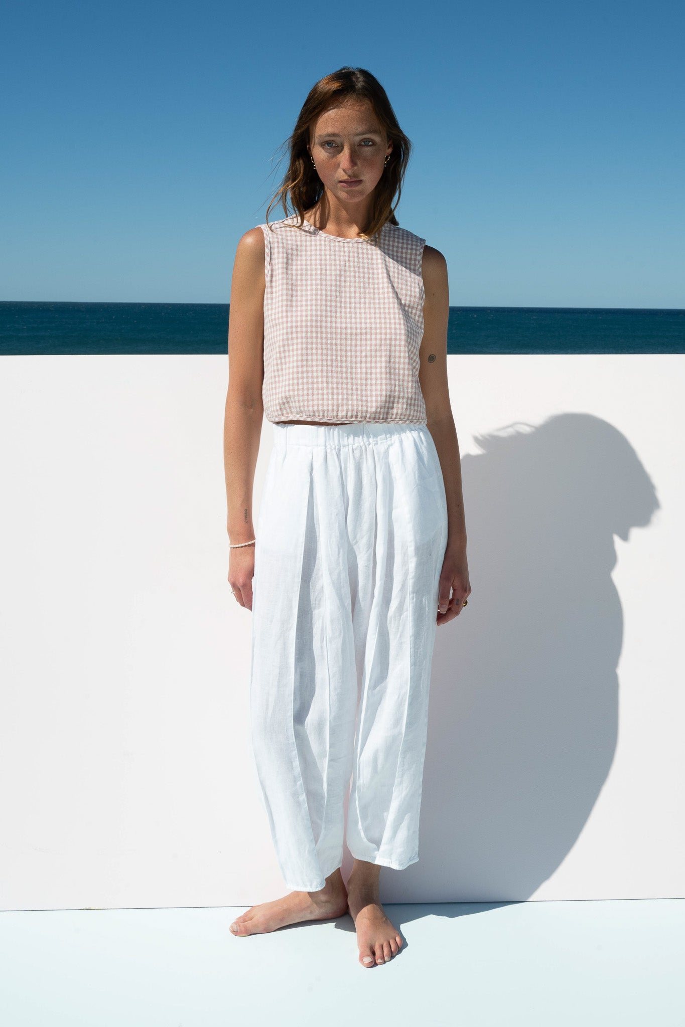 Front view of The Boy & I Box Crop Top in blush gingham cotton with white wall and ocean backdrop 