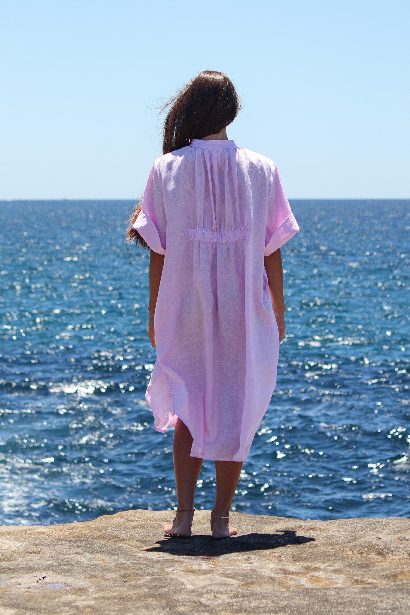Back view of the Cabana Dress in bonbon pink showing the gathered detail as the model looks out to sea