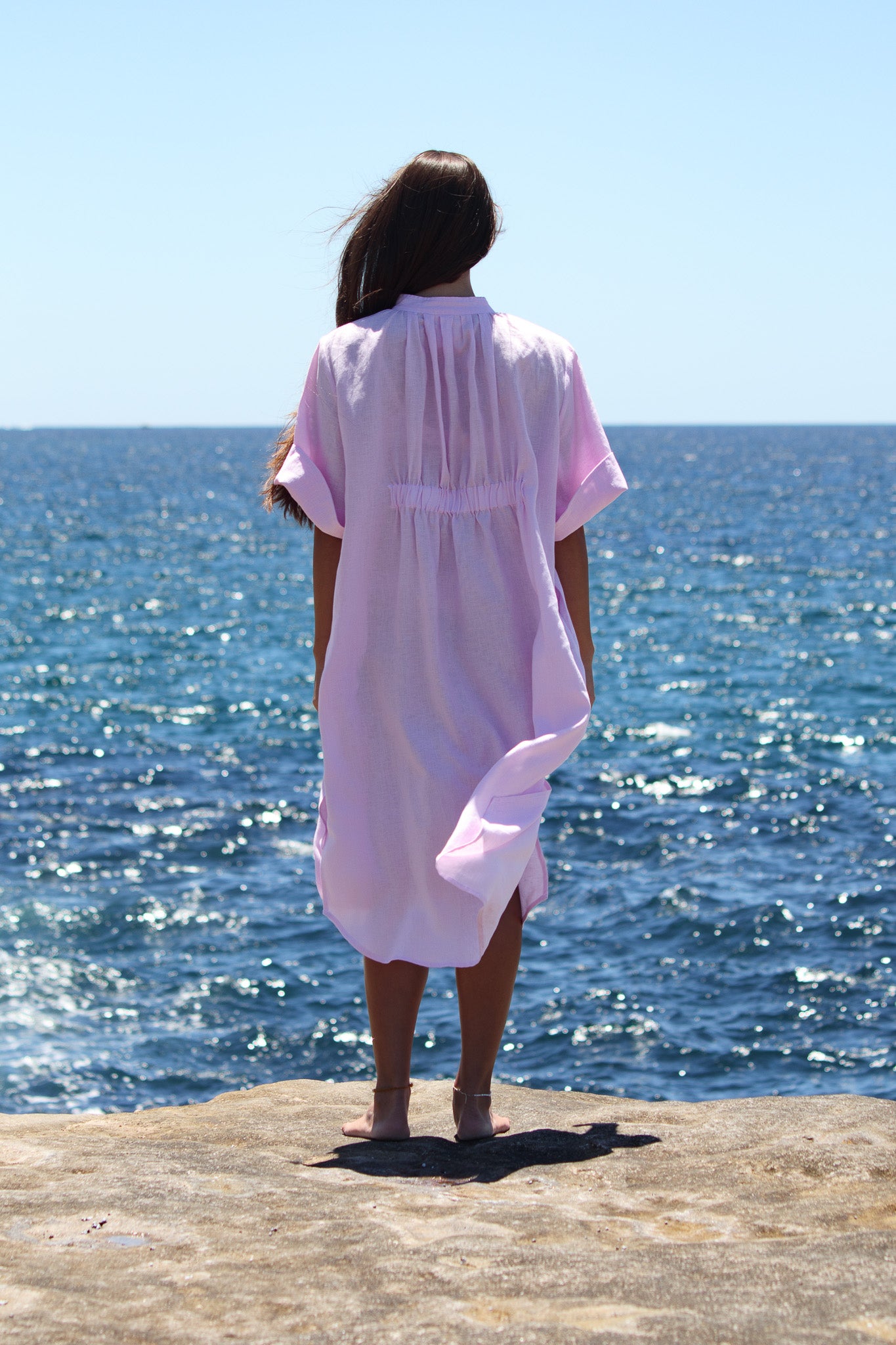 Model facing the ocean in the Cabana Dress in bonbon pink with the fabric moving in the breeze