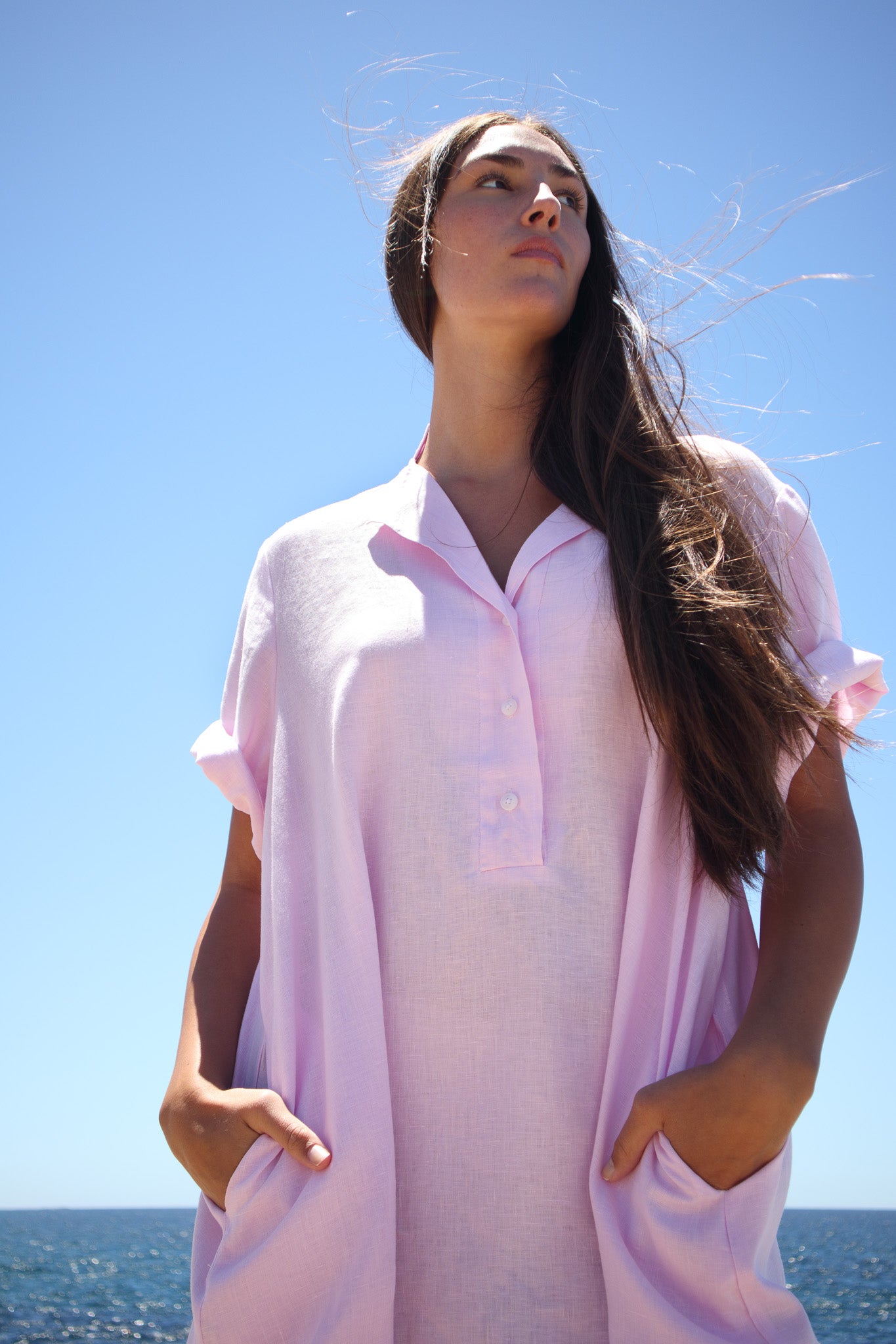 Model looking upward wearing the Cabana Dress in bonbon pink with the ocean and blue sky behind