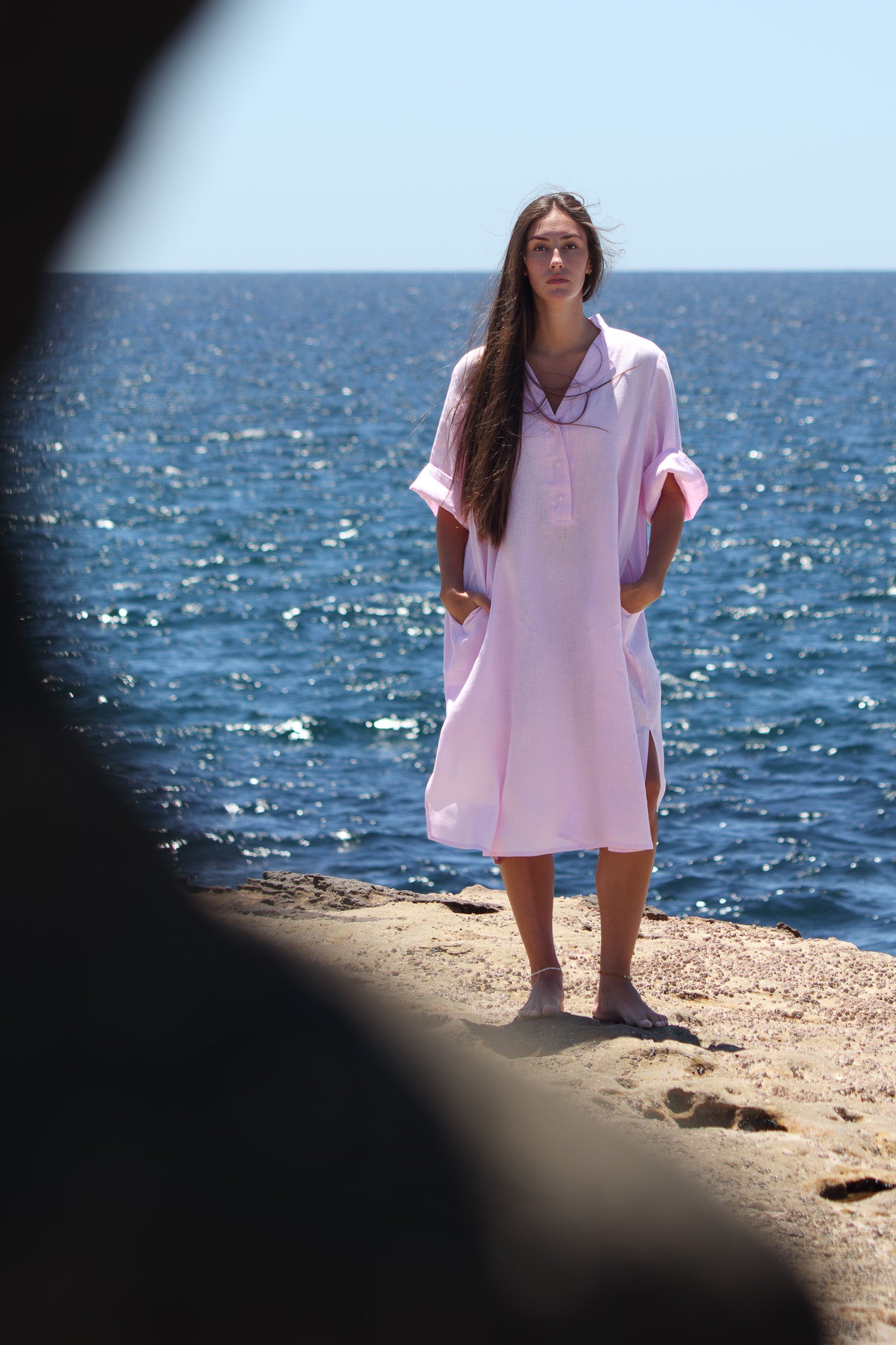 Model in the bonbon pink Cabana Dress framed by foreground rock with the ocean behind her