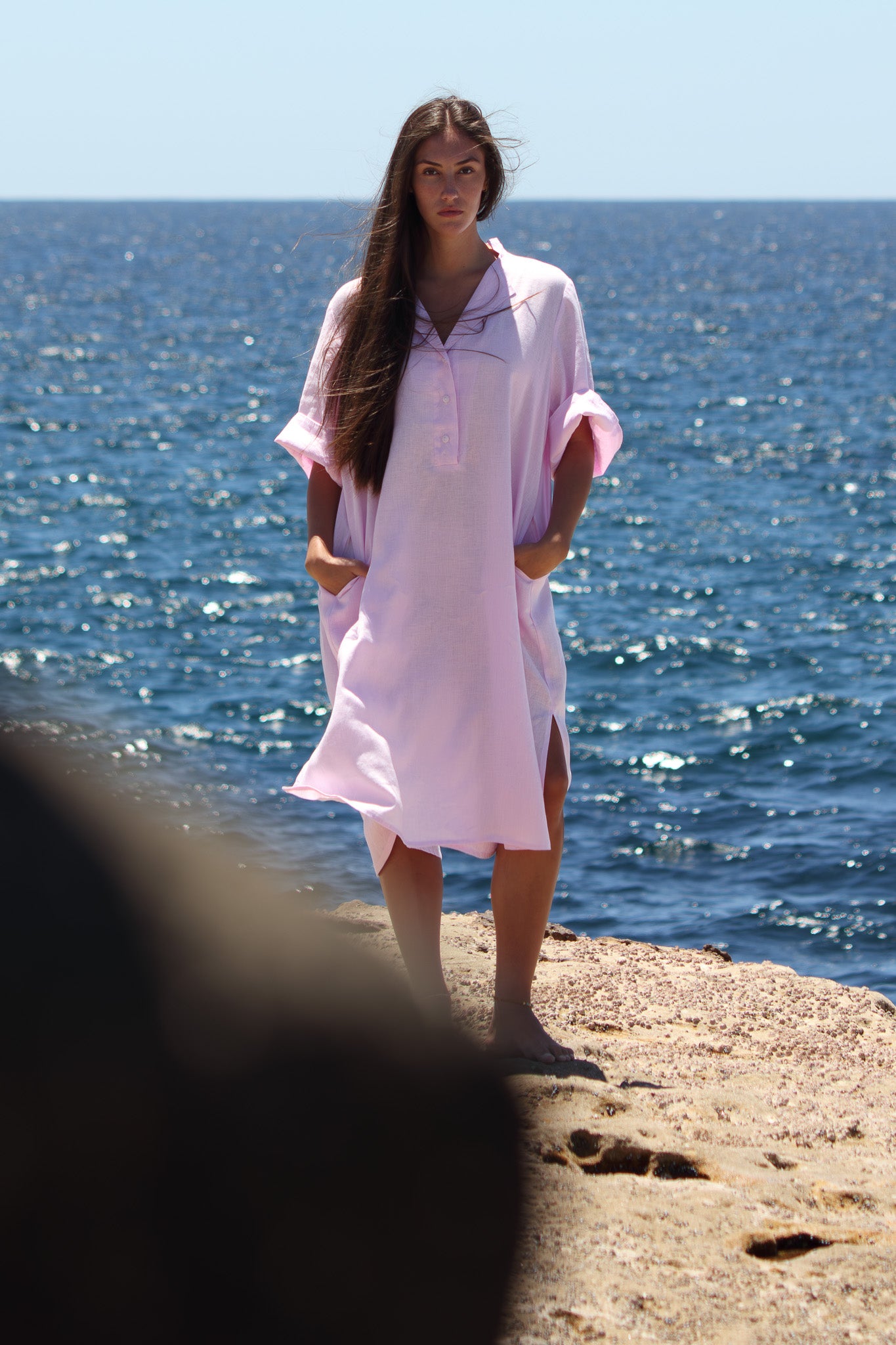 Model wearing the Cabana Dress in bonbon pink standing on coastal rock with hands in pockets