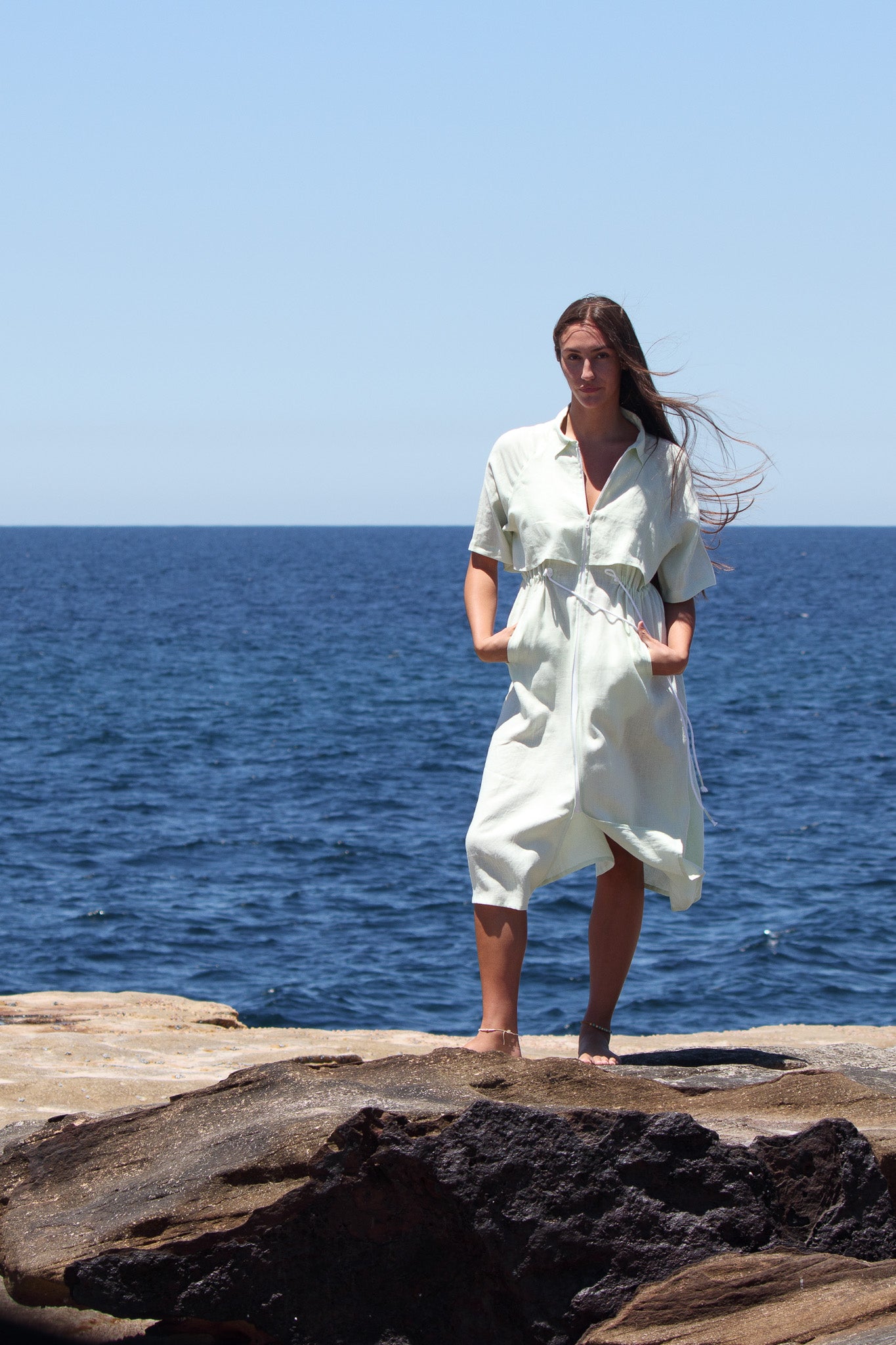 Model wearing the Iconica Dress in pistachio linen, standing full-length with the ocean behind, showcasing the relaxed drape and structured collar.