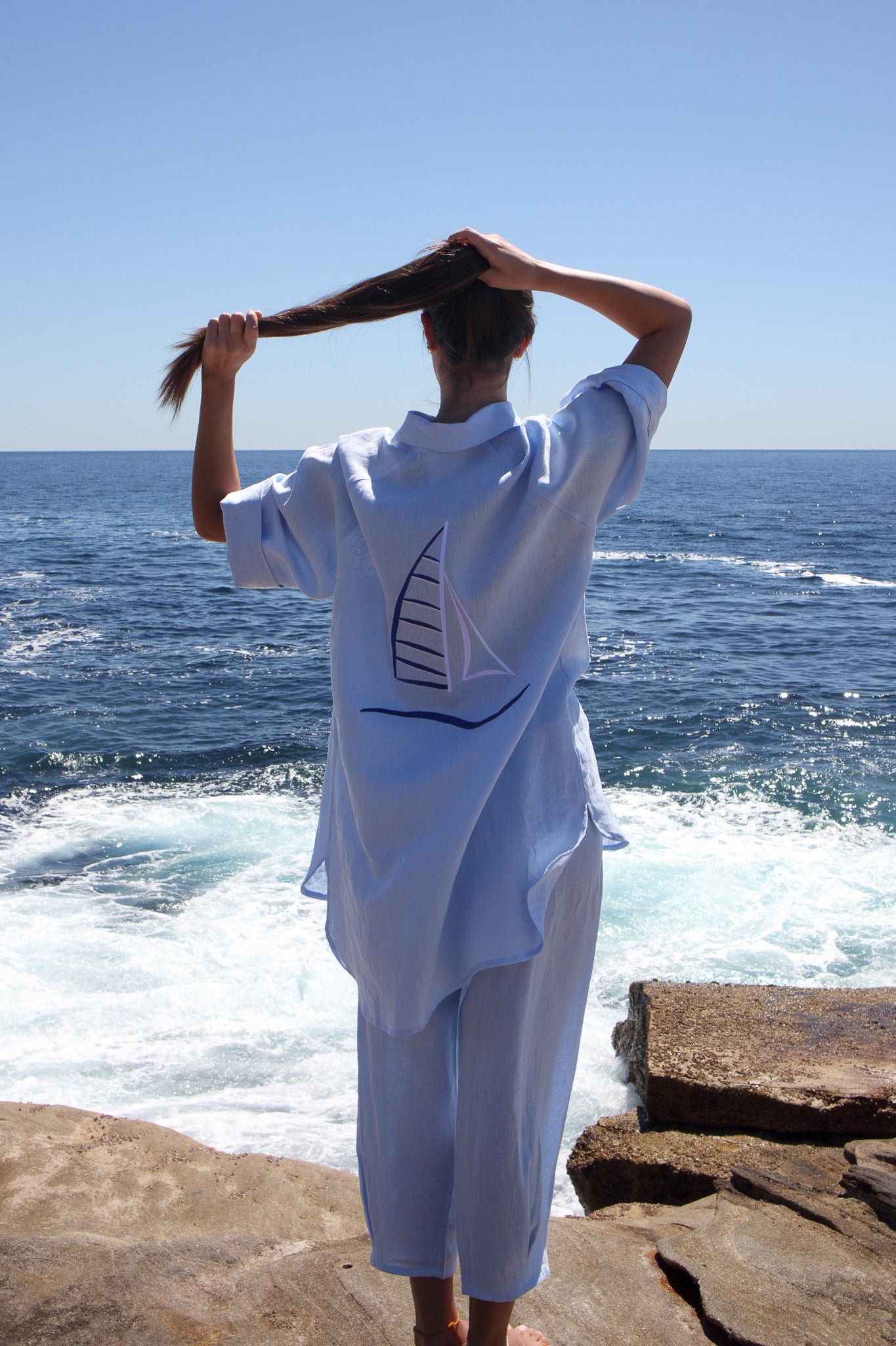 Back view of the Oceanic Shirt in sail boat showing embroidered sail design as model holds her hair by the water