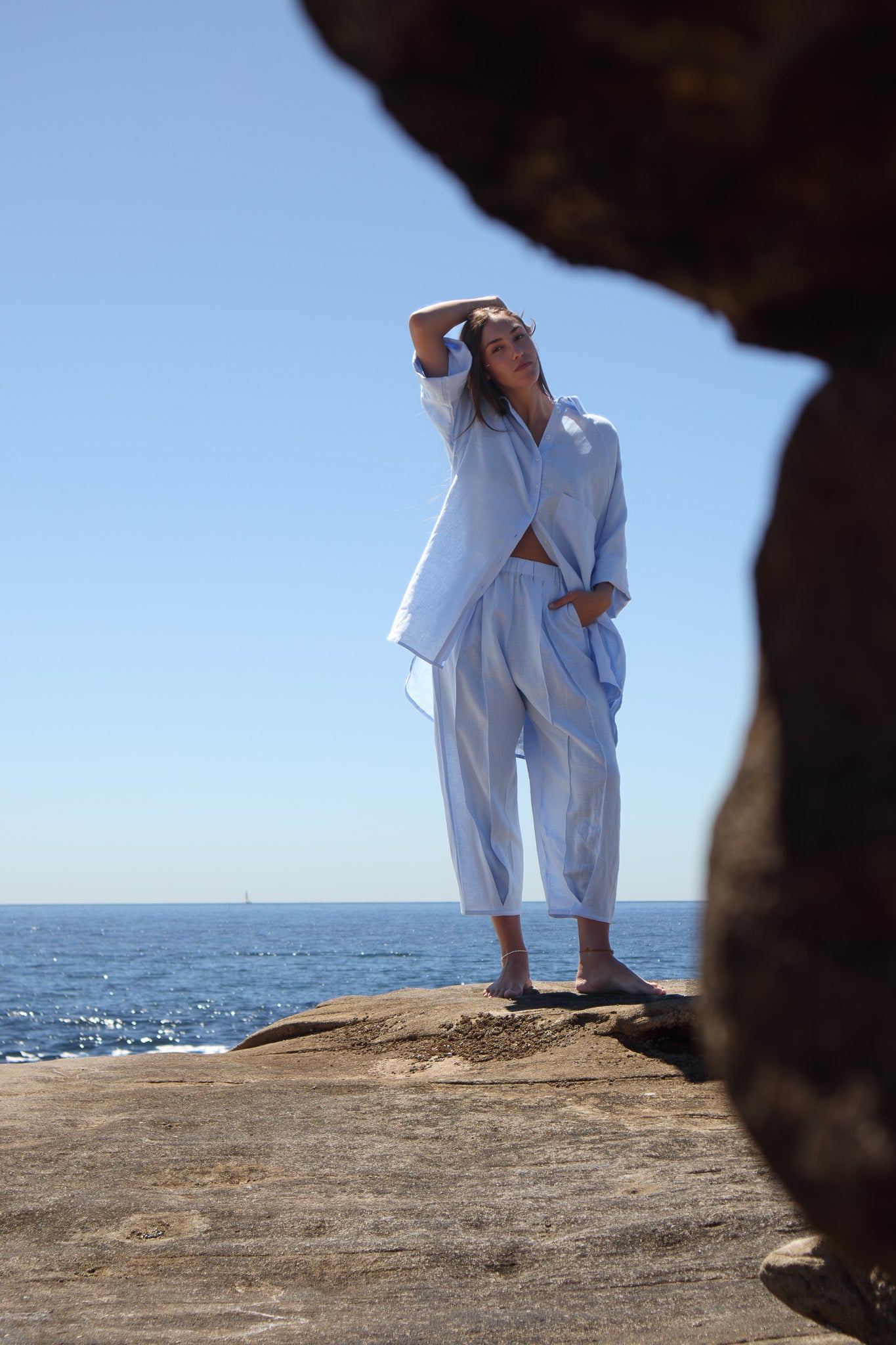 Model wearing the Oceanic Shirt in sail boat with matching pants standing on rocks by the ocean framed by foreground rock