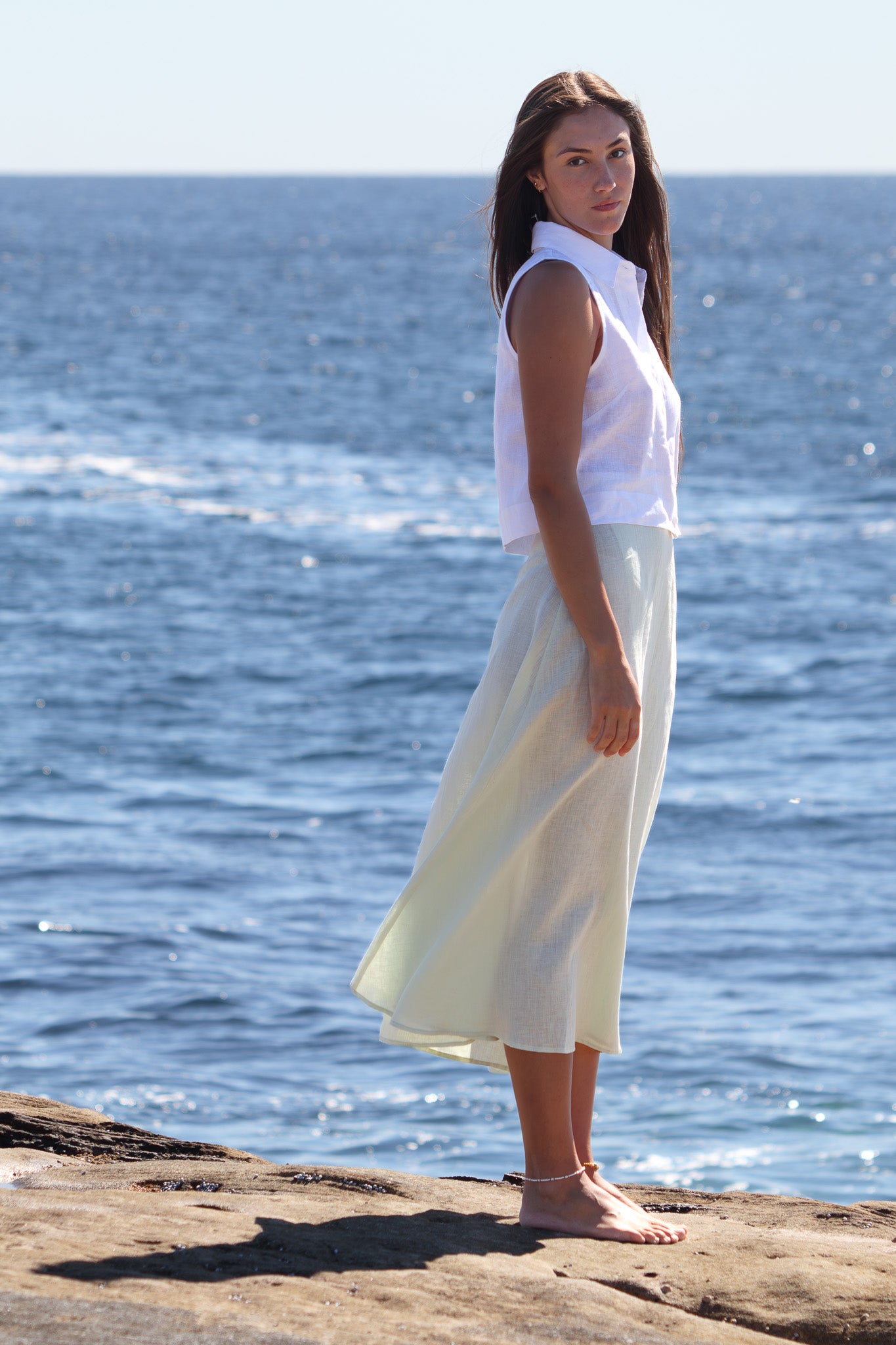Model wearing the PEPE Top in white linen with a pale linen skirt, photographed in soft natural light beside the sea.