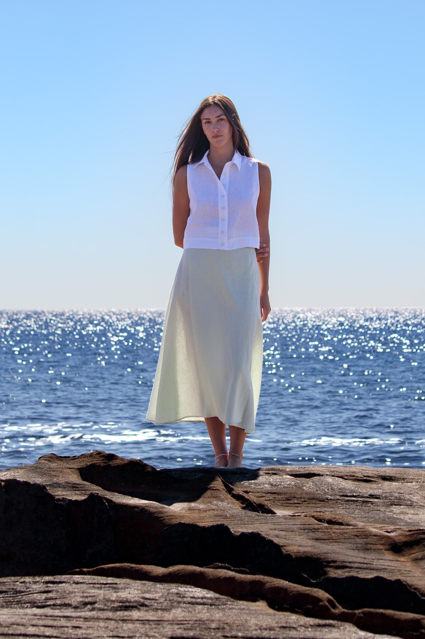 Full-length image of the model in the white PEPE Top and soft linen skirt, standing on the rocks with sparkling ocean behind.