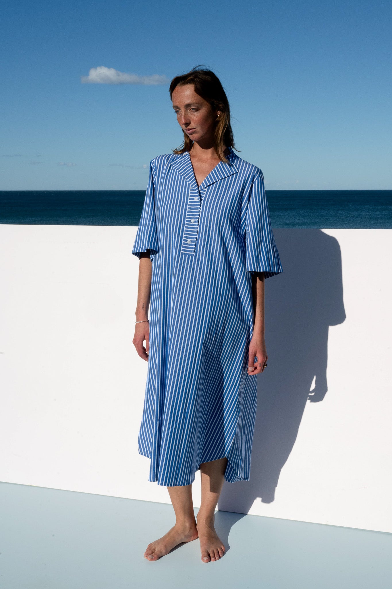 Full front view of the Cabana Dress in blue stripe cotton, styled barefoot against a coastal backdrop.