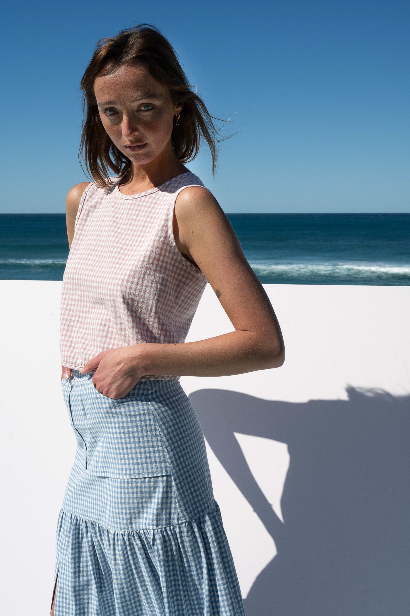 Angled portrait of model in Cancan Skirt blue gingham and blush crop top with ocean in background.