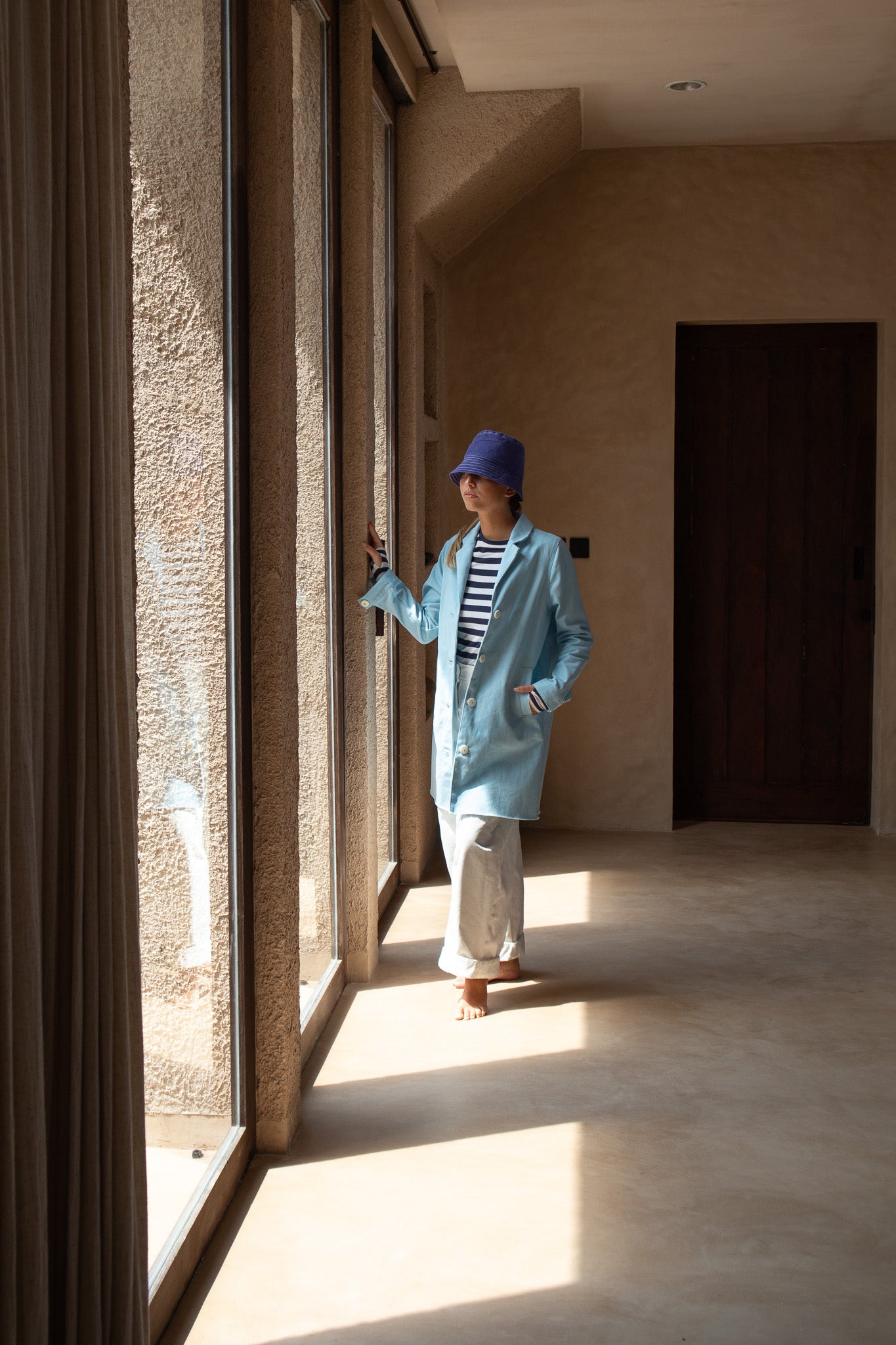 Model wearing the Hunter Coat in Light Sky Blue cotton twill suede, gazing out of sunlit windows.