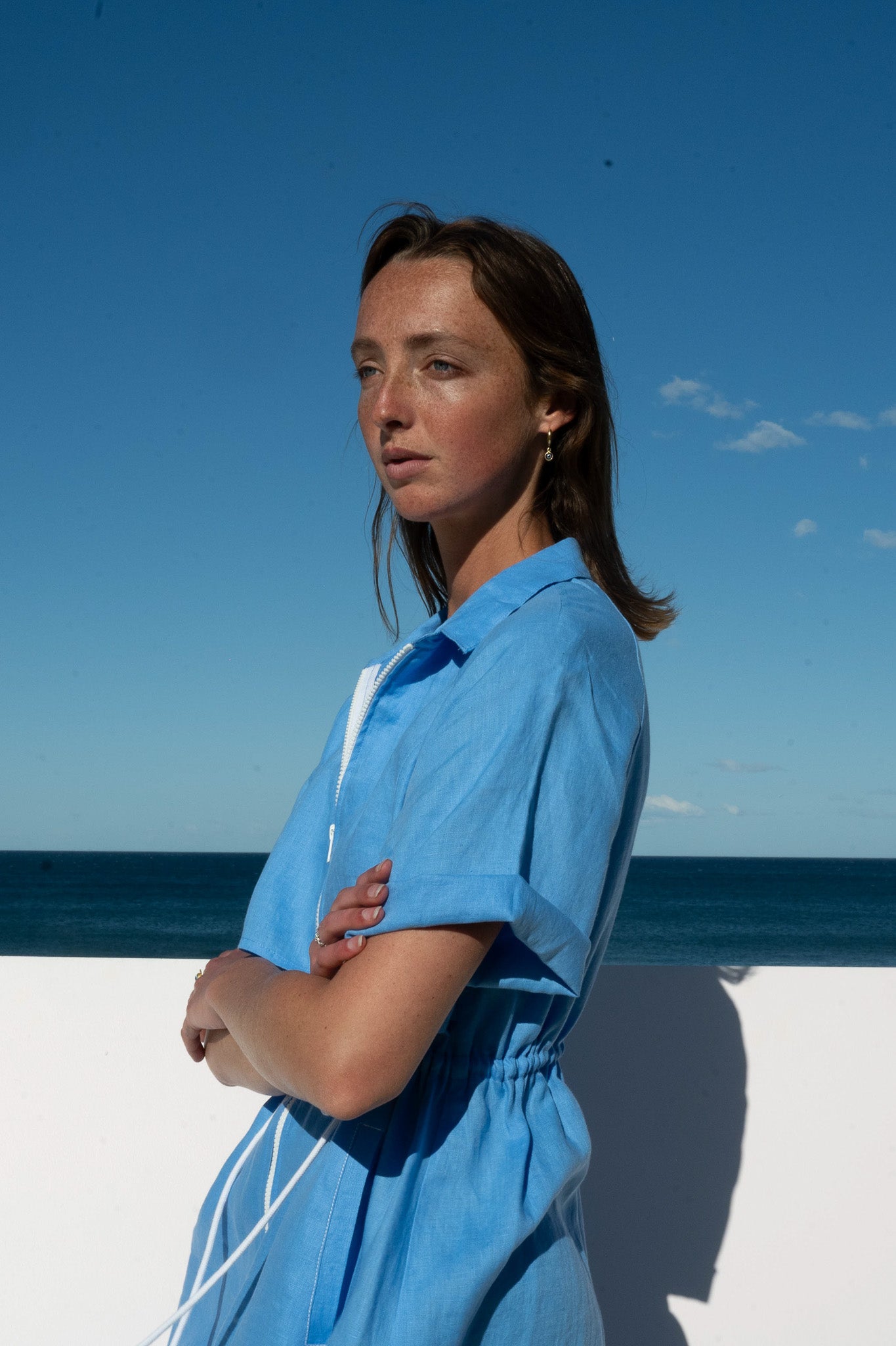 model wearing blue linen iconica dress with are folded and ocean and sky in the background 