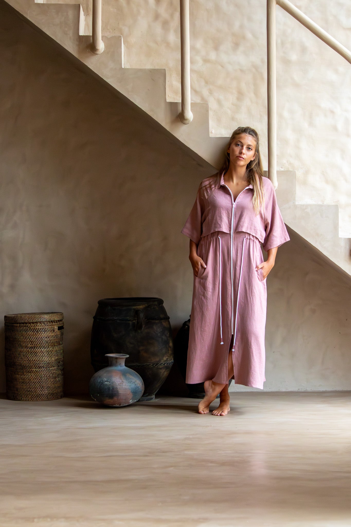 Full view of Iconica Dress in dusty pink with a white zip and drawstring, styled barefoot under a staircase.