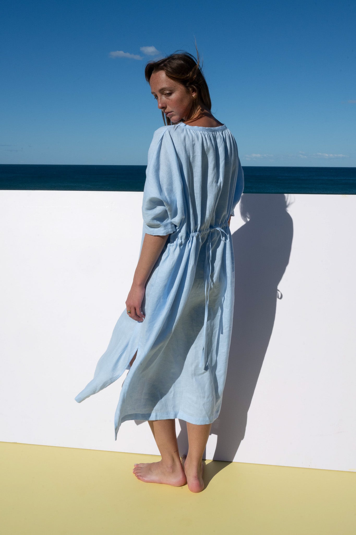 Back view of The Moments Dress in corn flour blue linen, showing gathered waist and tie fastening.