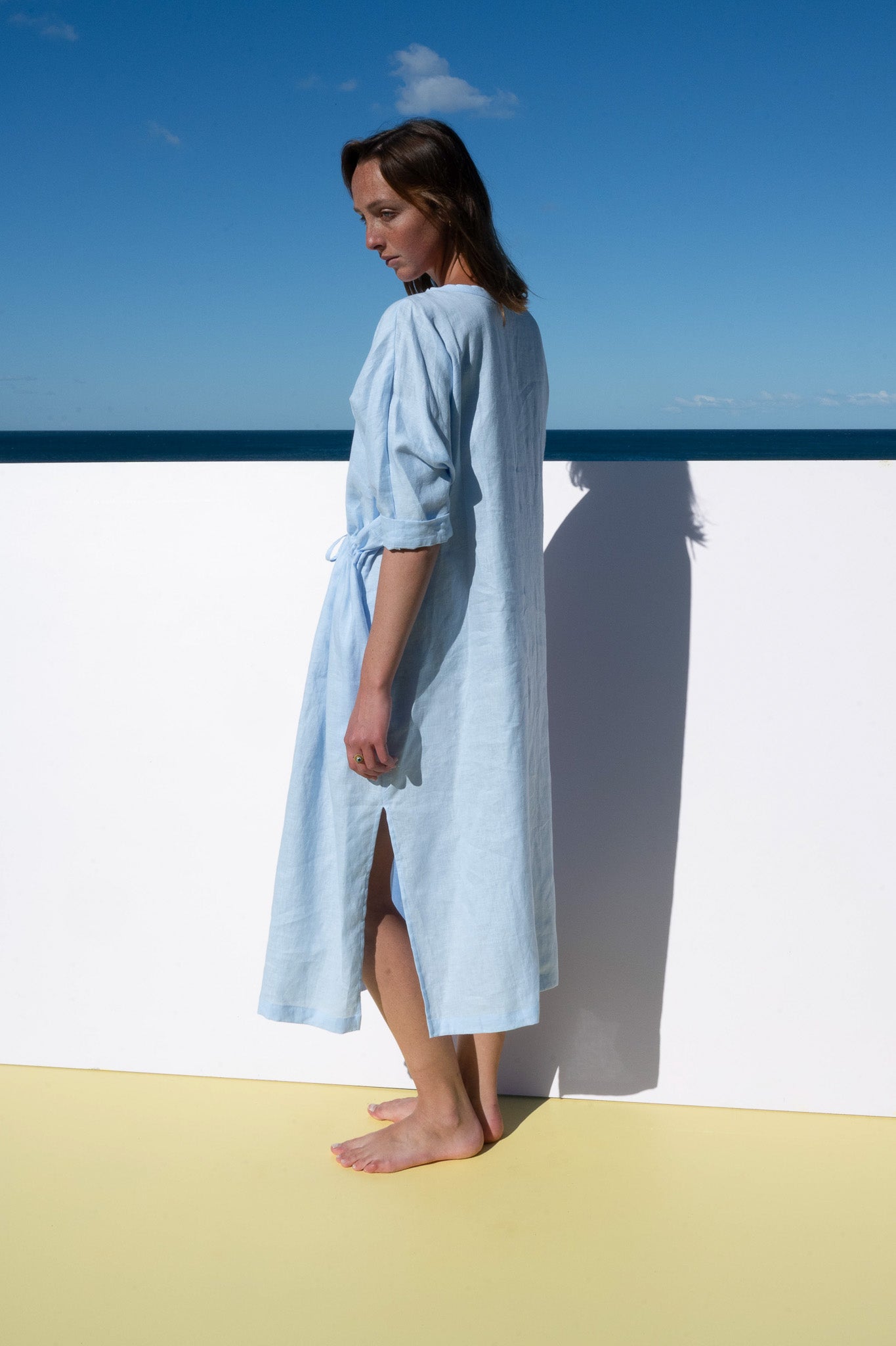 Model in The Moments Dress in corn flour blue linen, photographed side profile with tie detail visible.
