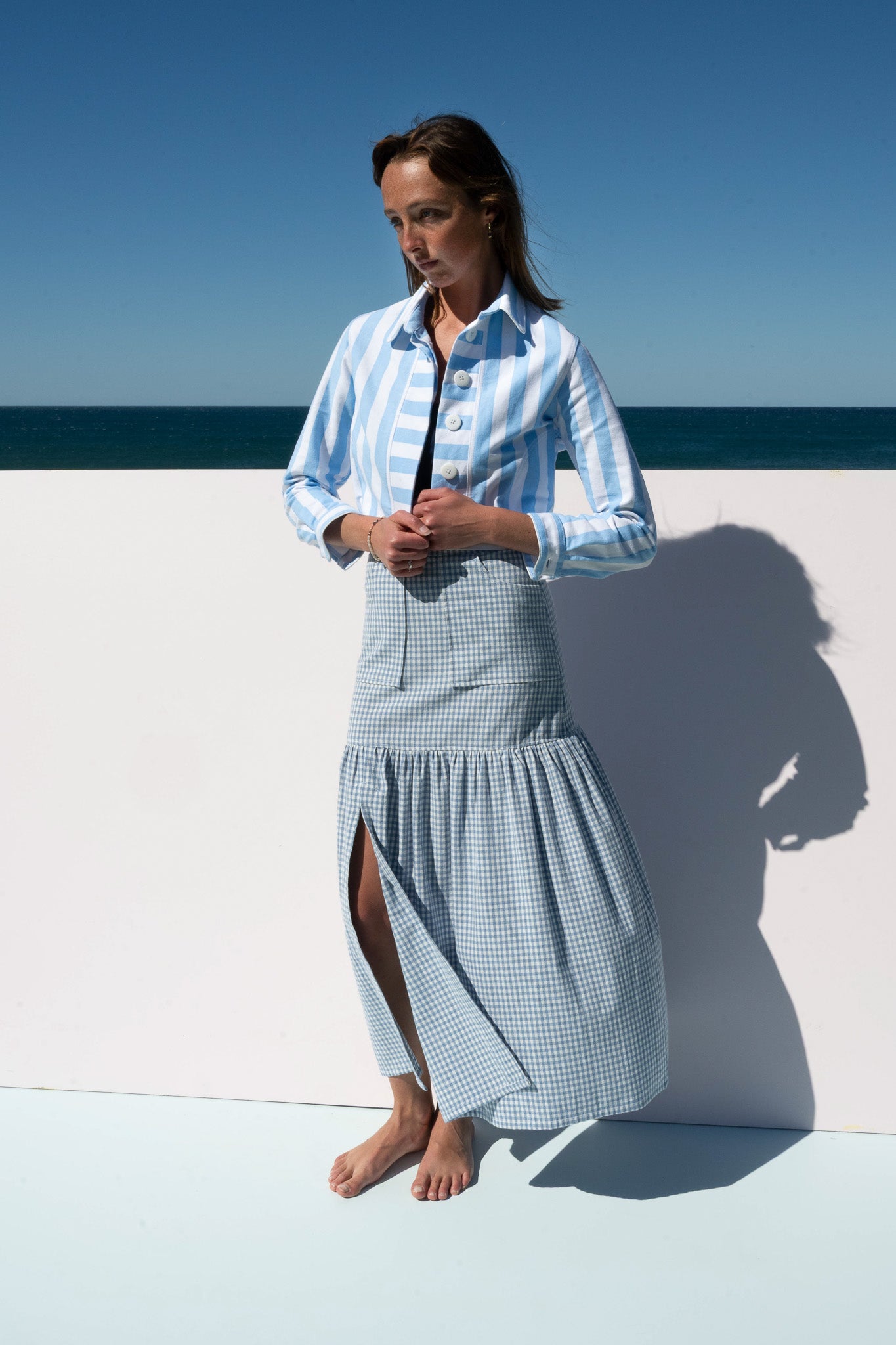 Model wearing The Boy & I Mon Petit Jacket in Saltwater Stripe styled with blue gingham skirt, seaside backdrop.