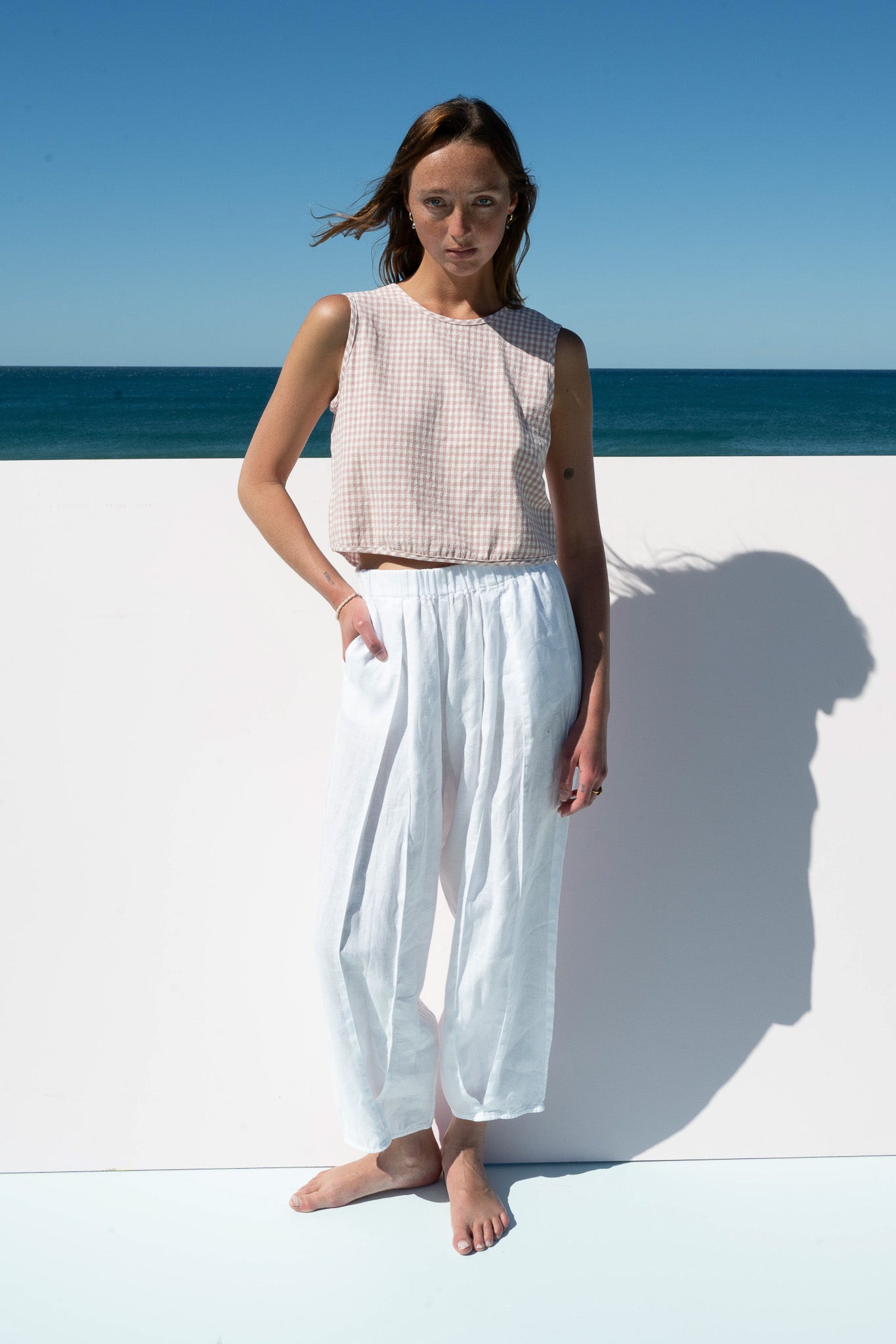 Model wearing Salt Pants in white linen with blush gingham crop top against beach backdrop.