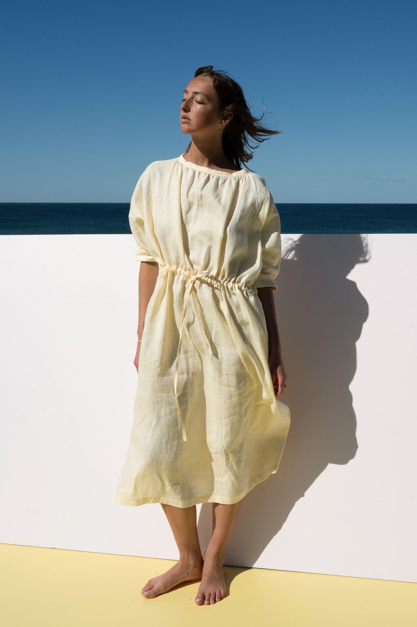 The Moments Dress in yellow linen – model standing in sunlight, showcasing loose fit and cinched waist