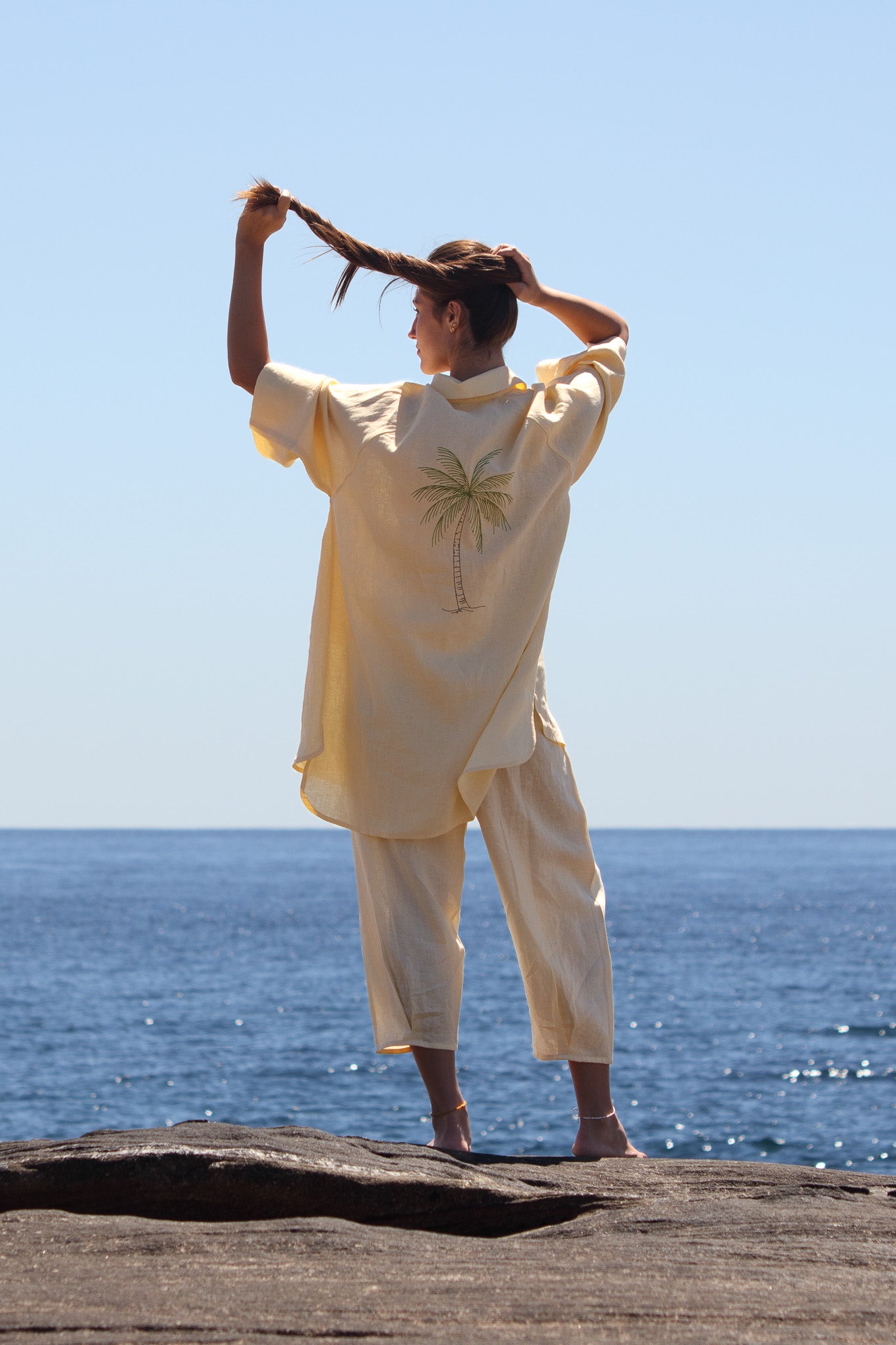 Back view of the butter yellow Oceanic Shirt Dress showing the embroidered palm tree and relaxed linen fit. person holding her hair