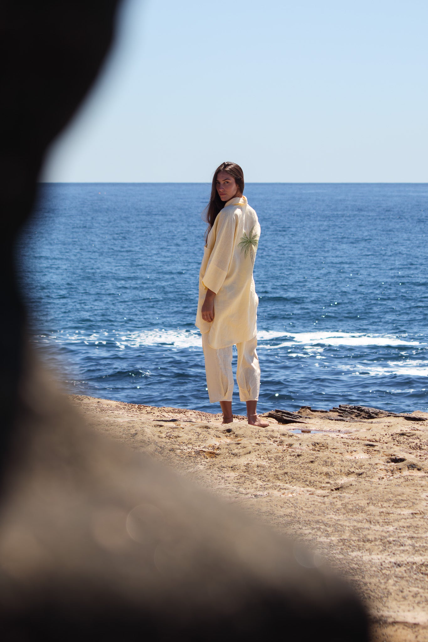 Oceanic Shirt Dress in butter yellow with palm tree embroidery on the back, worn by the water.